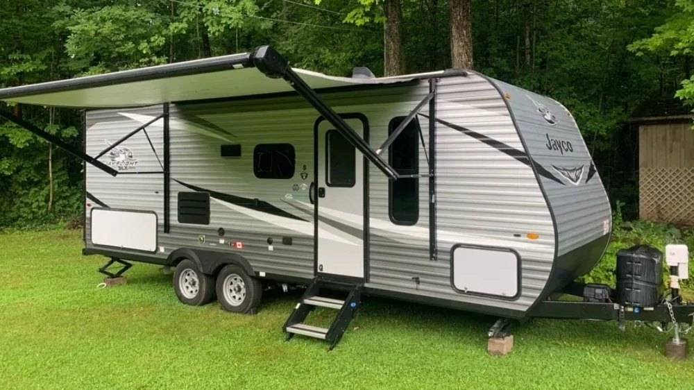 A travel trailer parked on grass with trees and a small shed in the background.