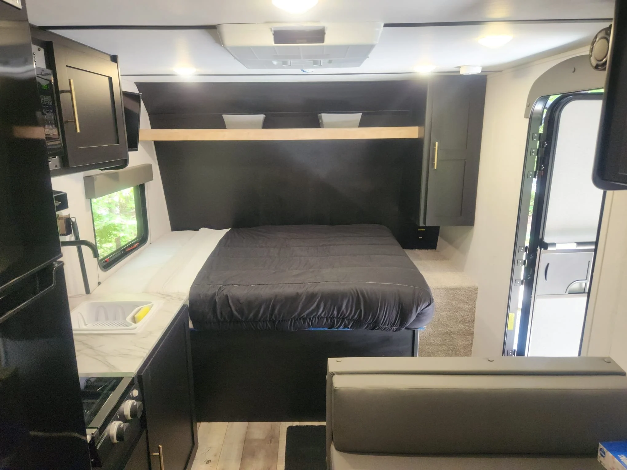 Interior of a modern camper or RV with a kitchenette, a bed with dark bedding, a window with greenery outside, and dark cabinetry with gold handles.