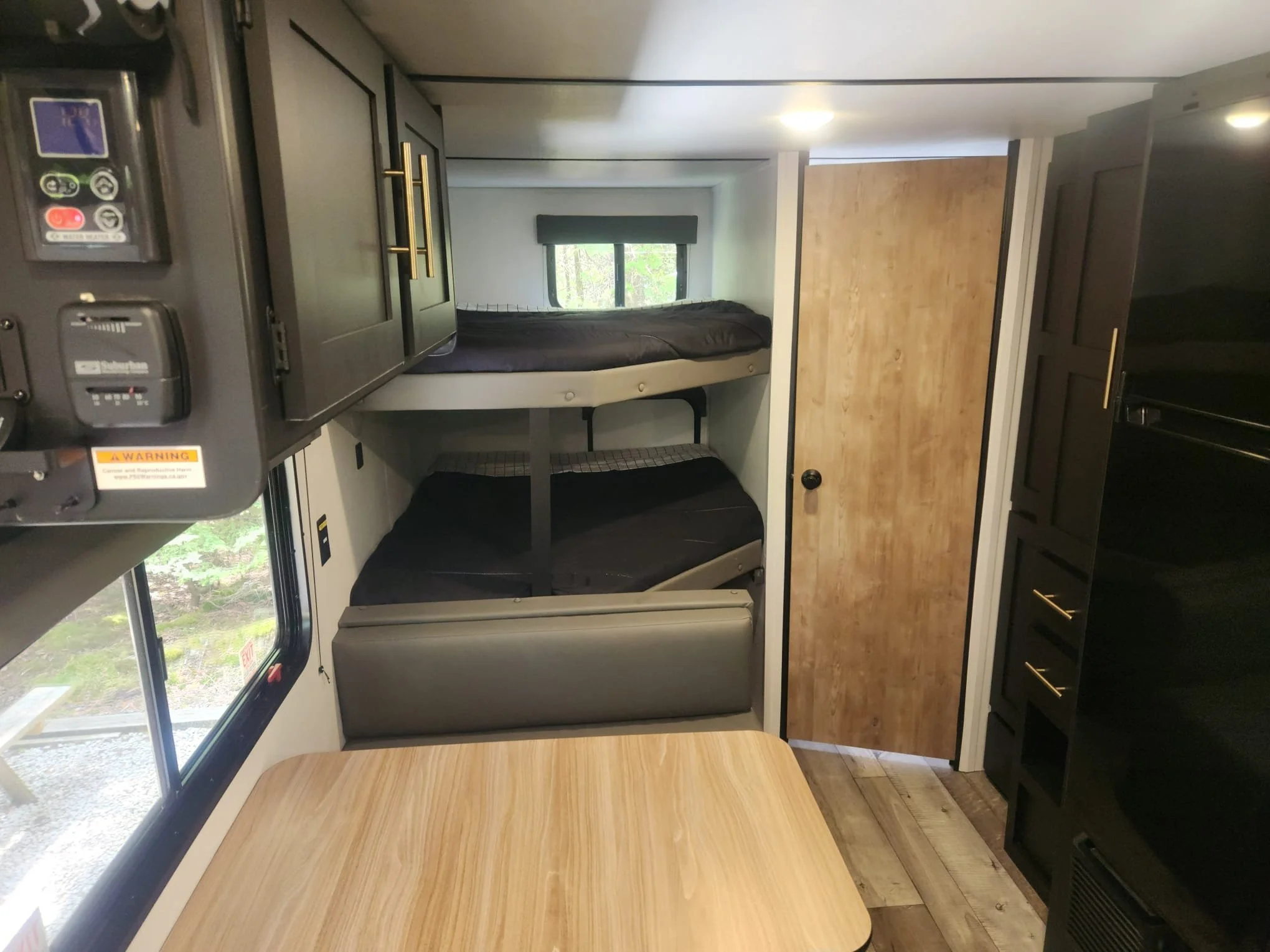 Interior of a compact camper or RV with a bed, window, black bedding, and wooden floor and cabinets.
