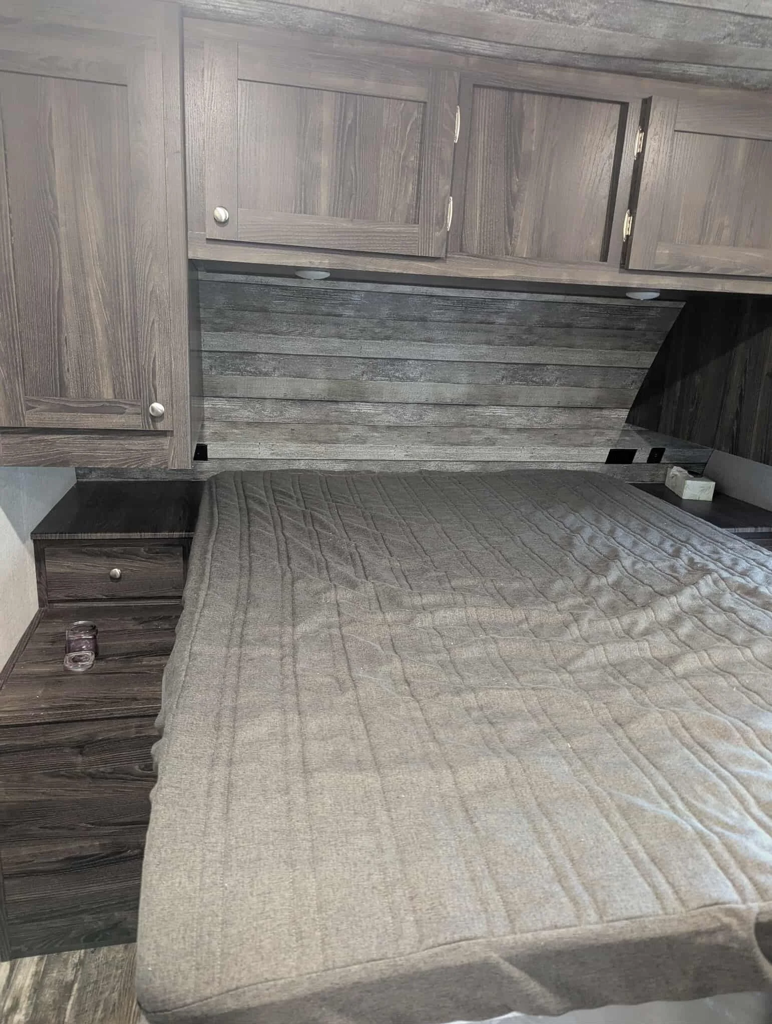 A bedroom with a wooden headboard, nightstand, and built-in cabinets, all in a wood grain finish, with a bed covered by a textured beige blanket.