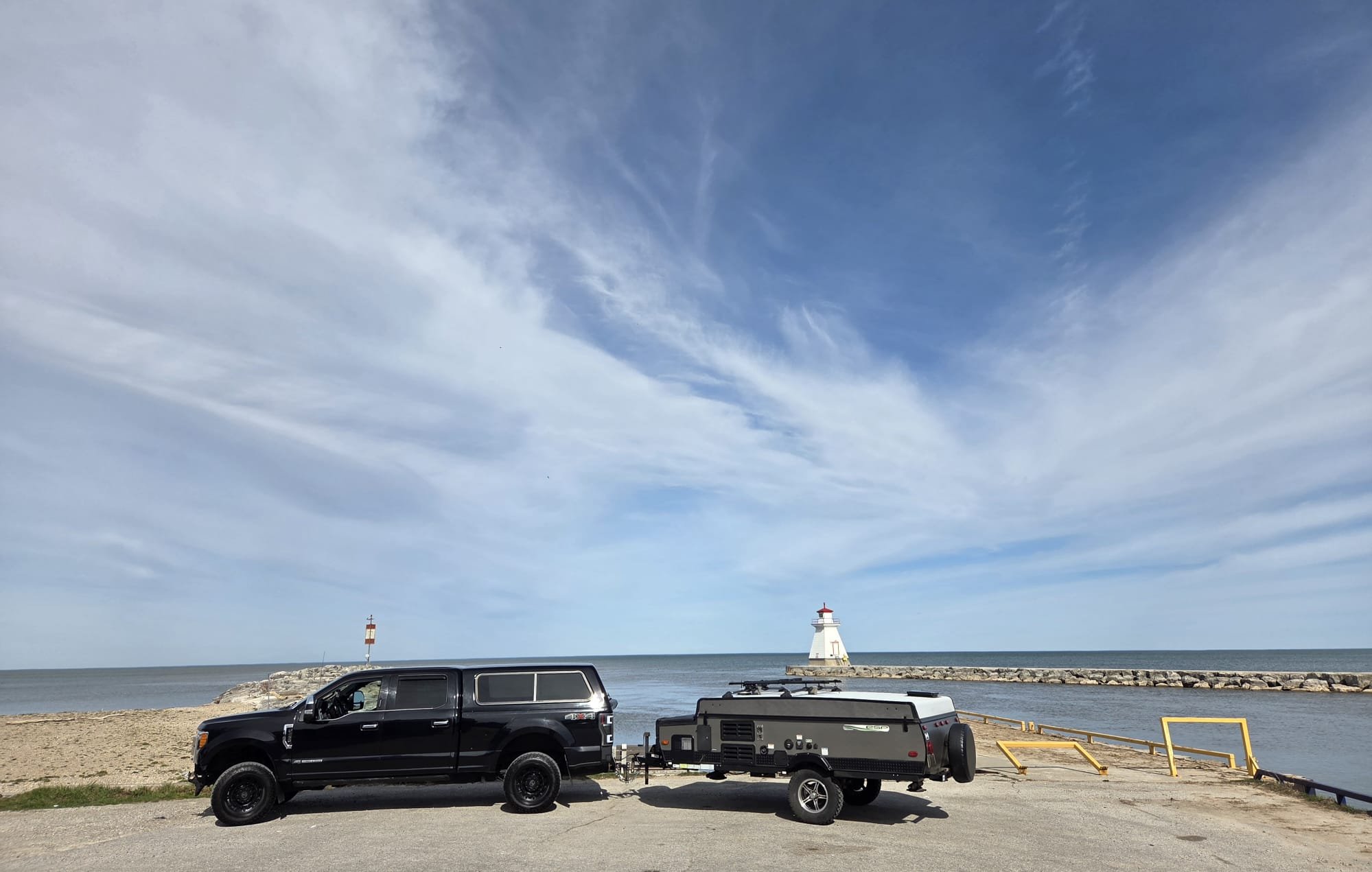 A picnic area by the water with a black truck, a portable generator, and a trailer, with a lighthouse and a breakwater in the background under a blue sky with scattered clouds.