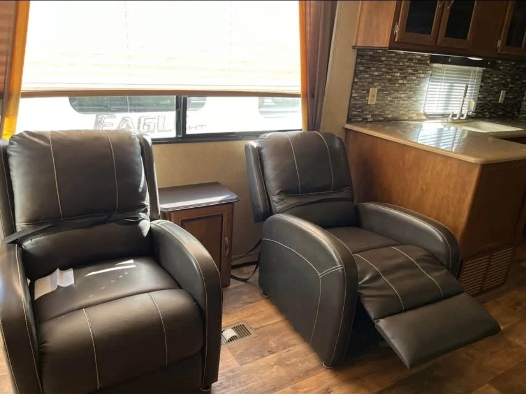 Two black leather recliner chairs in a living room near a window with blinds, a small wooden side table between them, and a kitchen with wooden cabinets, a granite countertop, and a tiled backsplash in the background.