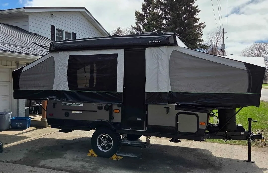 A black and white pop-up camper trailer parked on a residential driveway with houses and trees in the background.