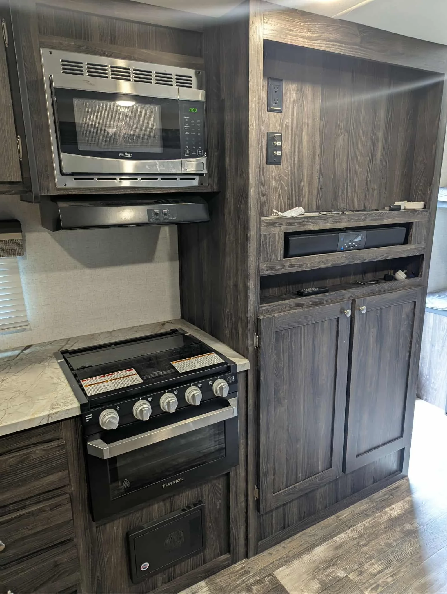 A kitchen with a stainless steel microwave above a stovetop and oven, with dark wood cabinets and a countertop below. To the right are dark wood panels with electrical outlets and an empty space or cabinet opening.