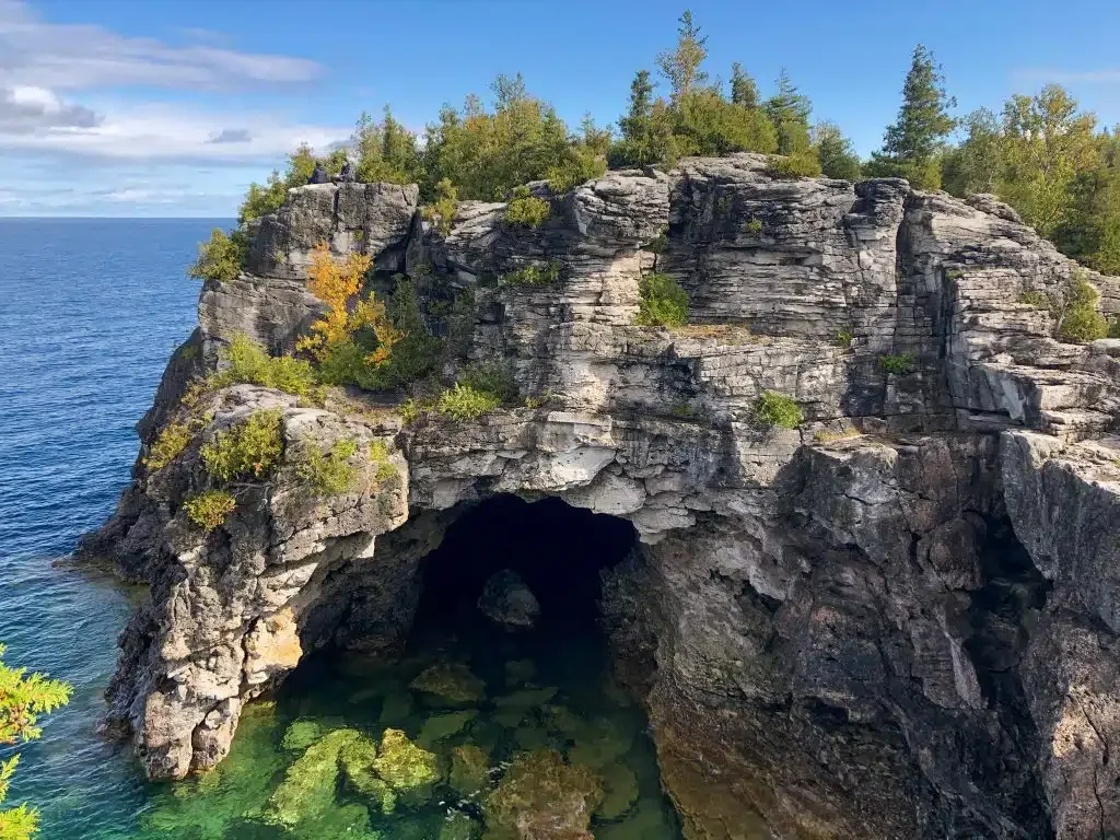 A rocky cliff with green trees and shrubs overlooking a body of water, featuring a natural large sea cave at its base.