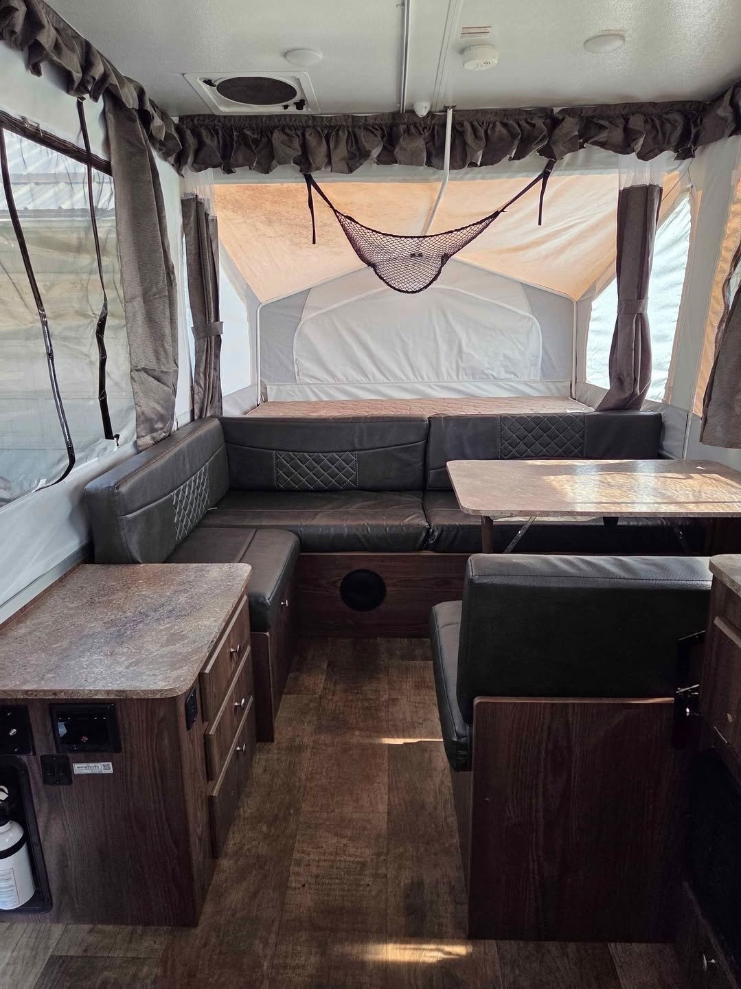 Interior of a camper or RV with a U-shaped black leather sofa, wooden tables, and windows with curtains. There is a small net hanging from the ceiling and some cabinets near the floor.