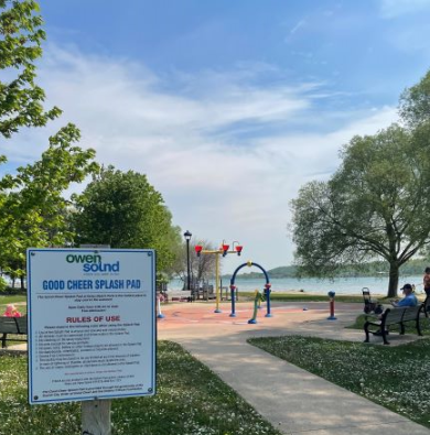 A splash pad at Owen Sound with water sprinklers, a lake in the background, and benches along the pathway.