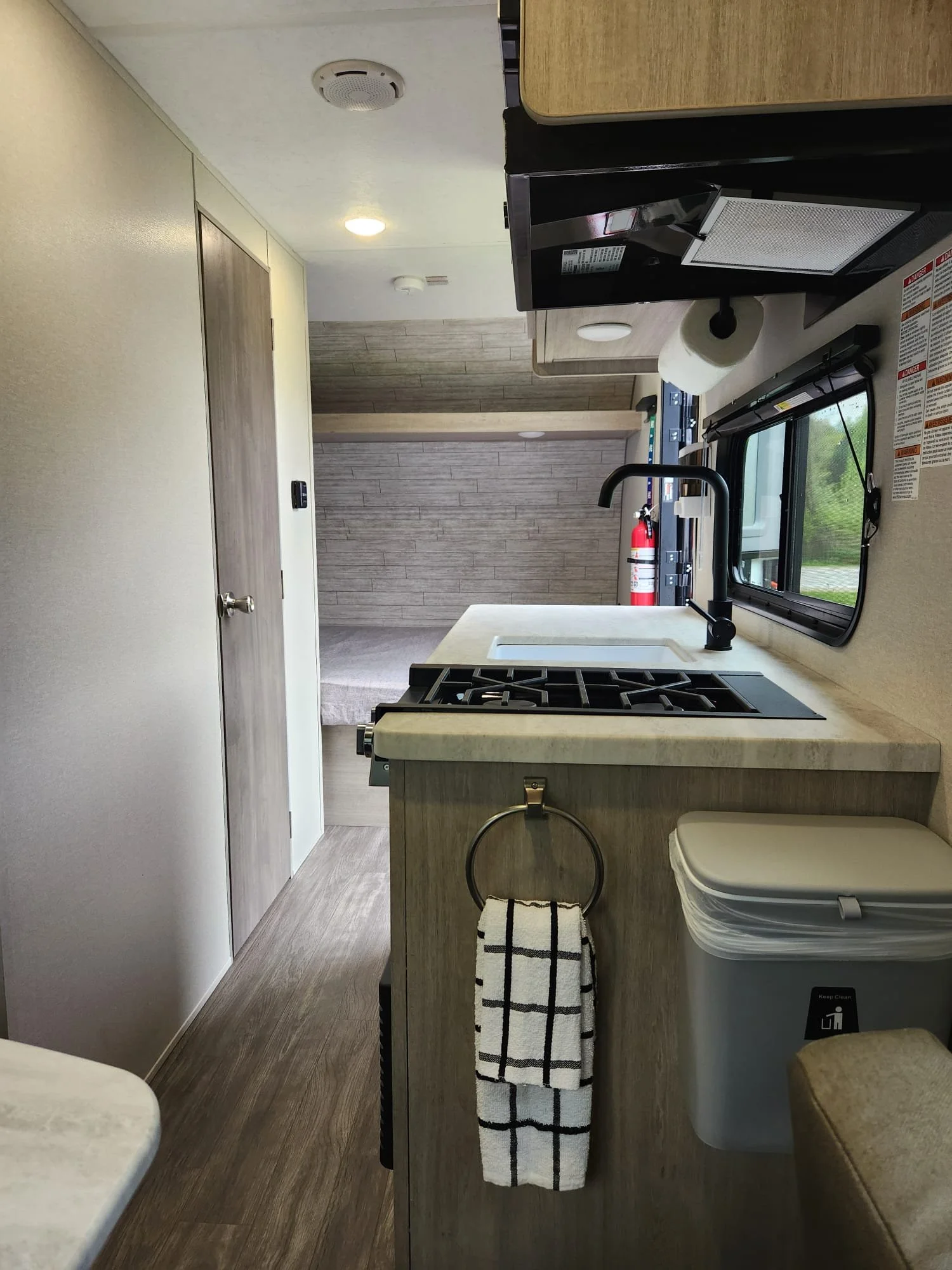 Interior of a compact camper kitchen with a stove, sink, window, and wooden cabinets. A fire extinguisher is mounted near the window, and a small trash can is on the counter.