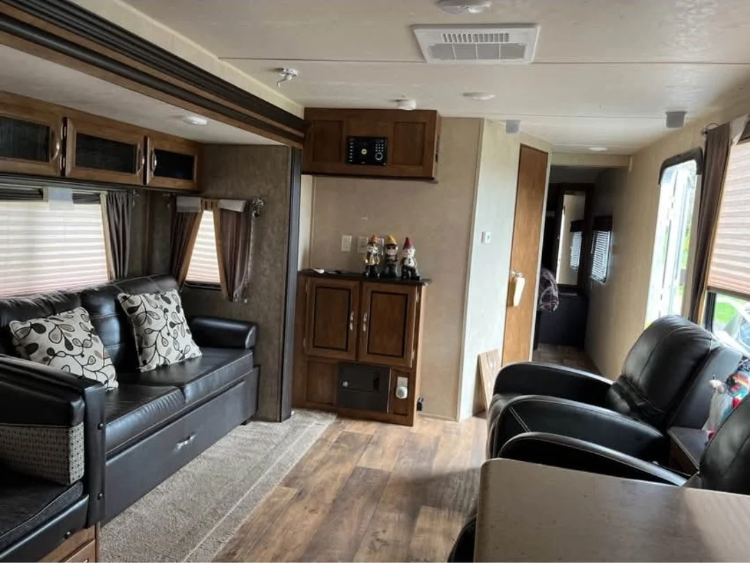 Living room inside a recreational vehicle with black leather couch, armchair, and wooden cabinets, decorated with Christmas figurines and bulbs, with windows dressed in curtains and blinds.