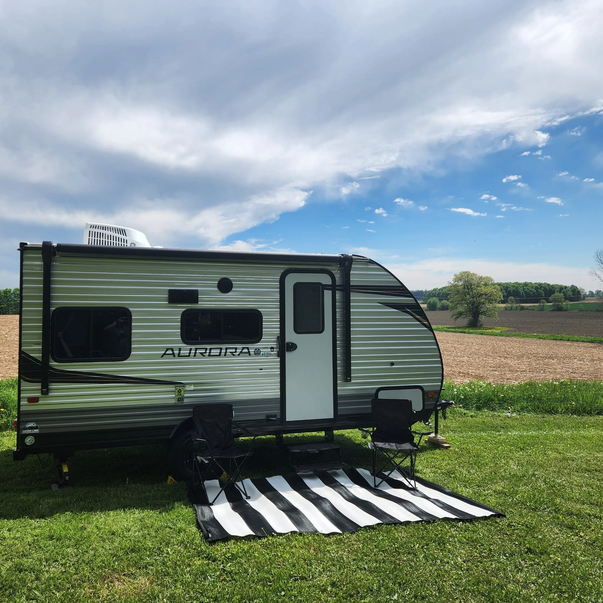 A travel trailer with the word 'Aurora' on the side, parked on a grassy area with a striped mat and outdoor chairs. The sky is partly cloudy with some blue visible and green trees in the background.
