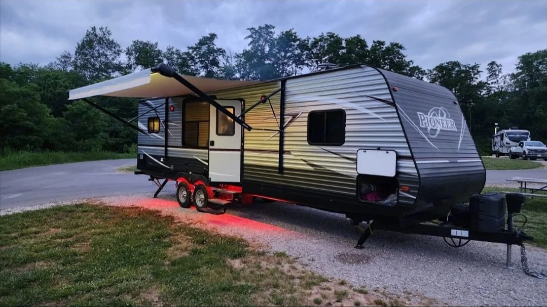 A Pioneer travel trailer with an extended awning and red underglow lights parked on a gravel site near a grassy area and a tree-lined horizon.