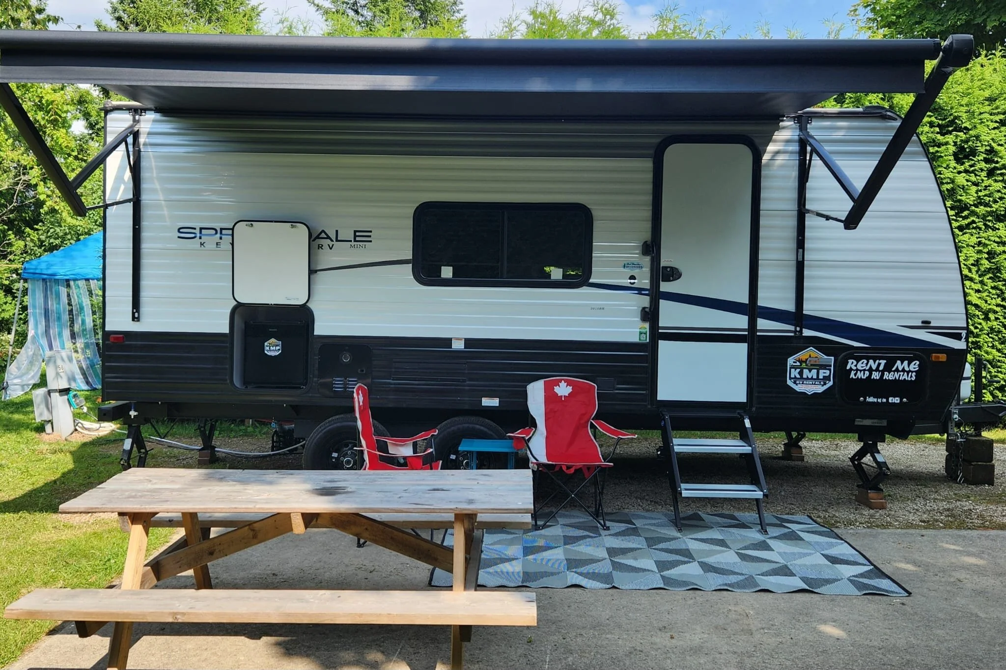 A travel trailer with a retractable awning, set up outdoors with a picnic table and chairs underneath a patterned rug, against a background of green trees and a blue sky.