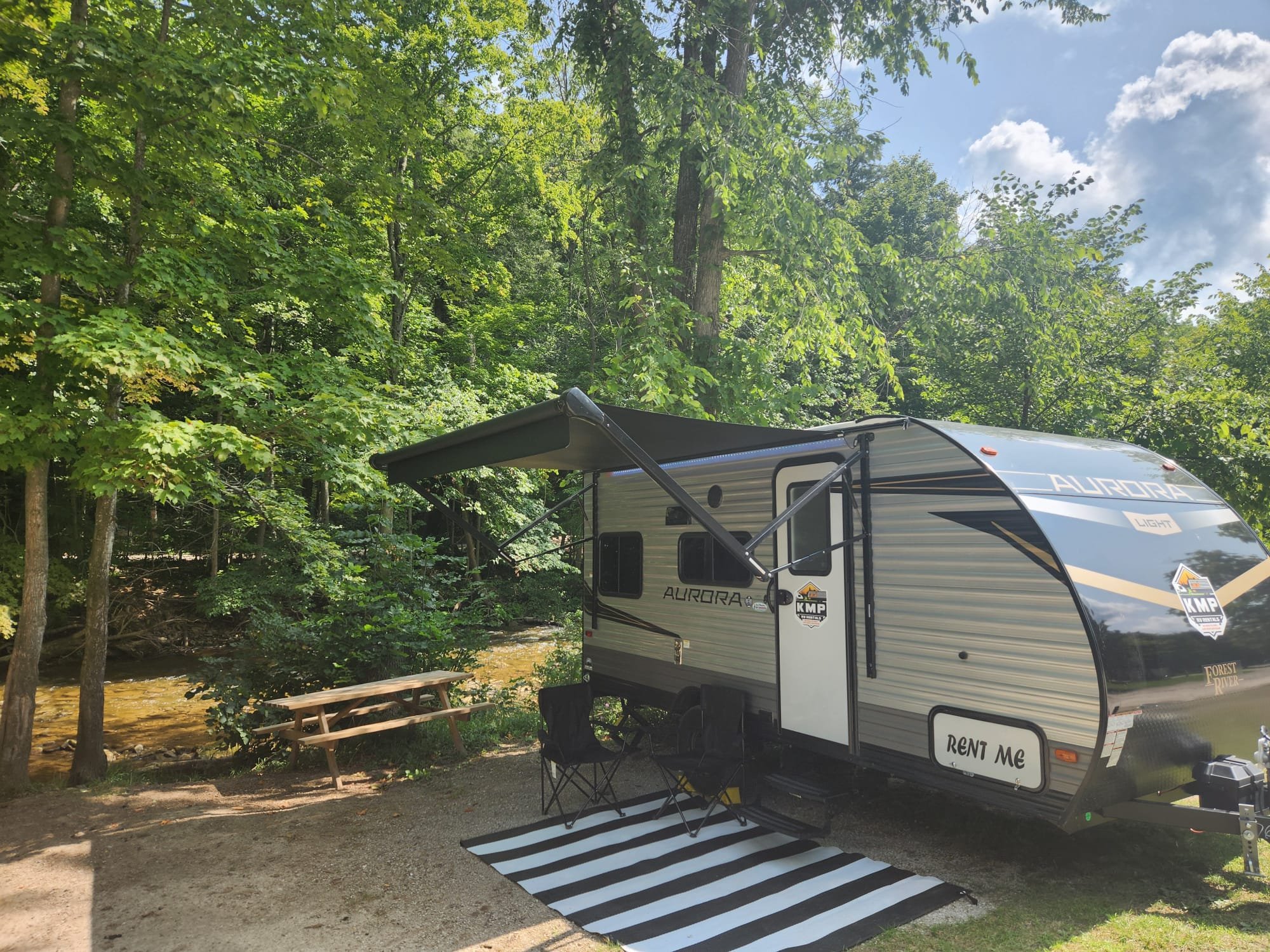 A travel trailer parked in a wooded area next to a river, with an outdoor picnic table and foldable chairs, under a digital striped outdoor mat, with trees and a partly cloudy sky in the background.