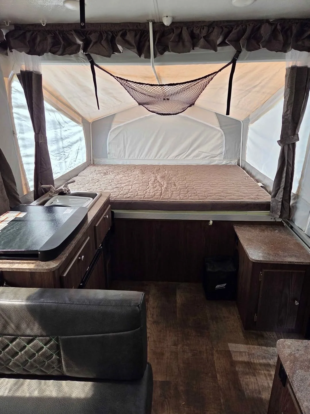 Interior of a small camper with a raised bed, a kitchenette with a sink, and a small table. The bed has a mattress, and there are curtains and windows on either side.