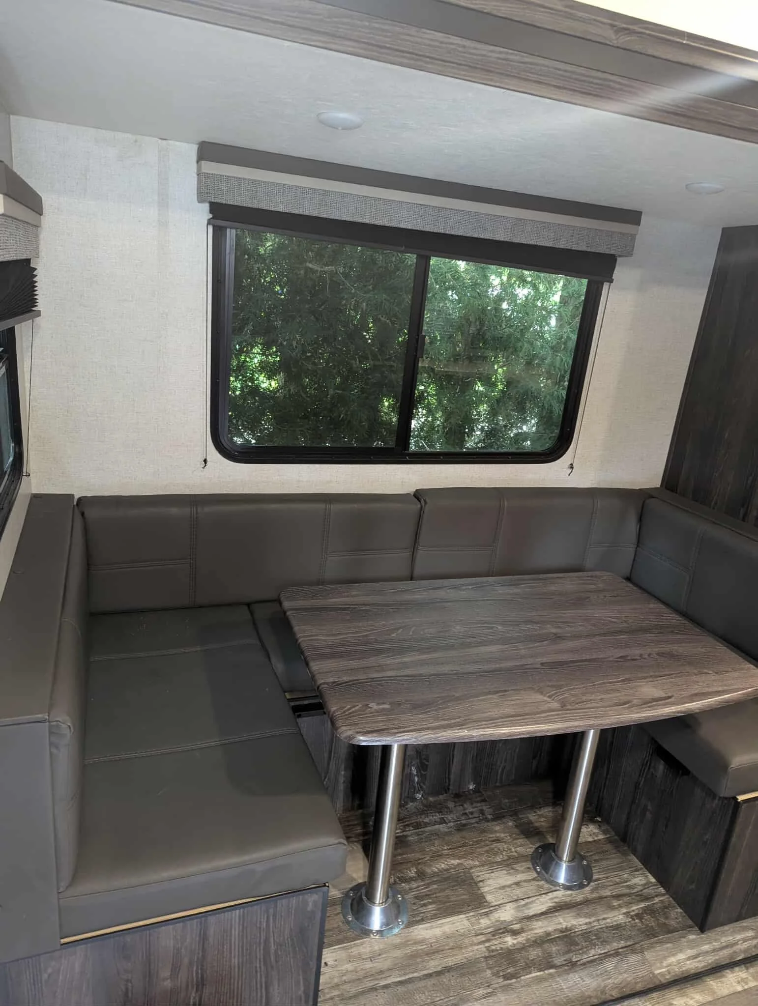 Interior of a recreational vehicle or camper with a large window showing outdoor trees, a seating area with gray L-shaped cushioned benches, a small wooden table with metal legs, and fabric window coverings.