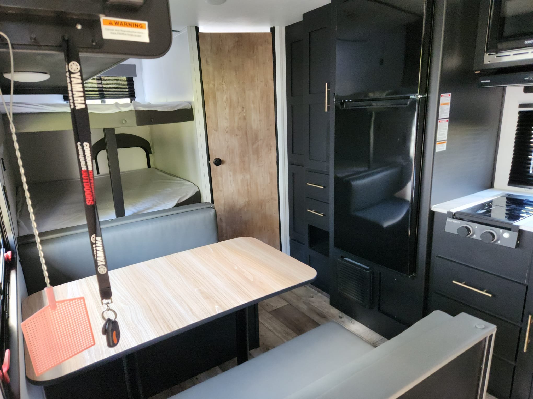Interior of a compact kitchen in a RV with black cabinets, a stove, a microwave, a small dining table, and bunk beds with white mattresses.