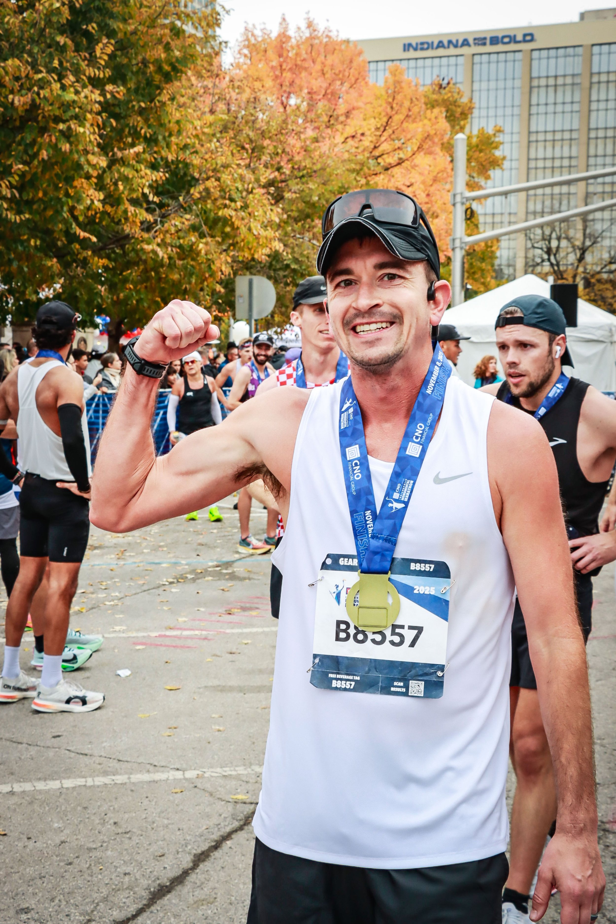 A smiling man in a white running tank top and black cap, showing his flexed arm, at a marathon event with other runners and an autumn tree in the background.