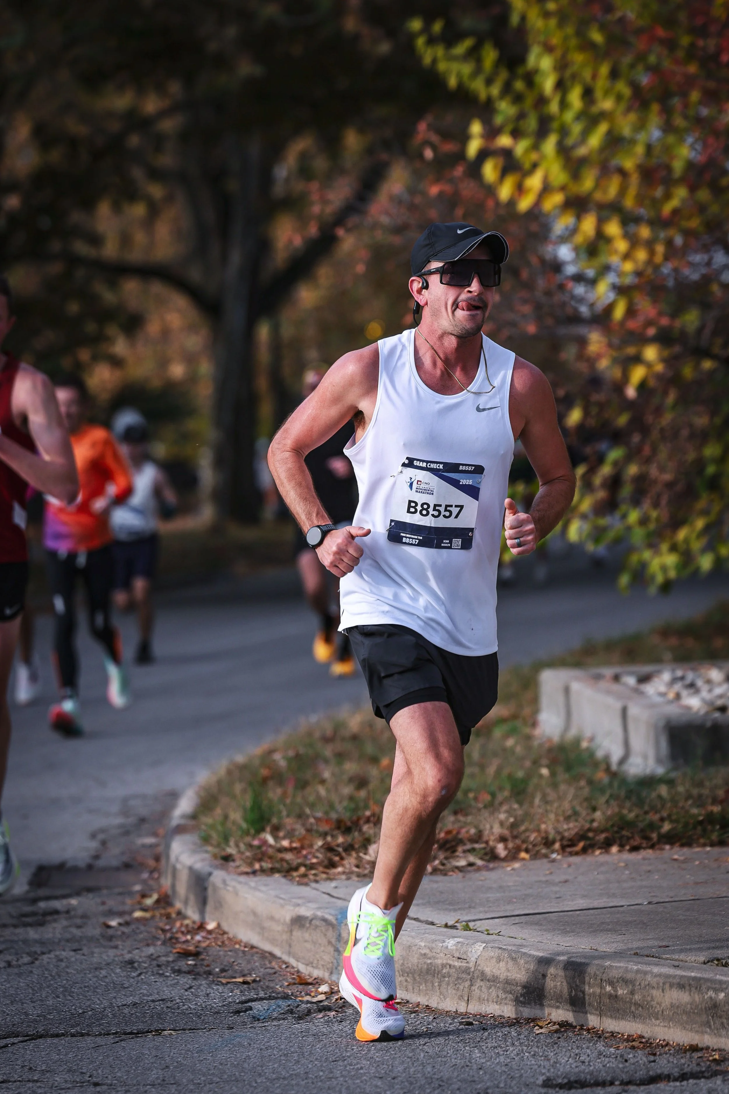 A man running in a marathon wearing a white sleeveless shirt, black shorts, sunglasses, a sports watch, and a black cap, with other runners and trees with autumn leaves in the background.