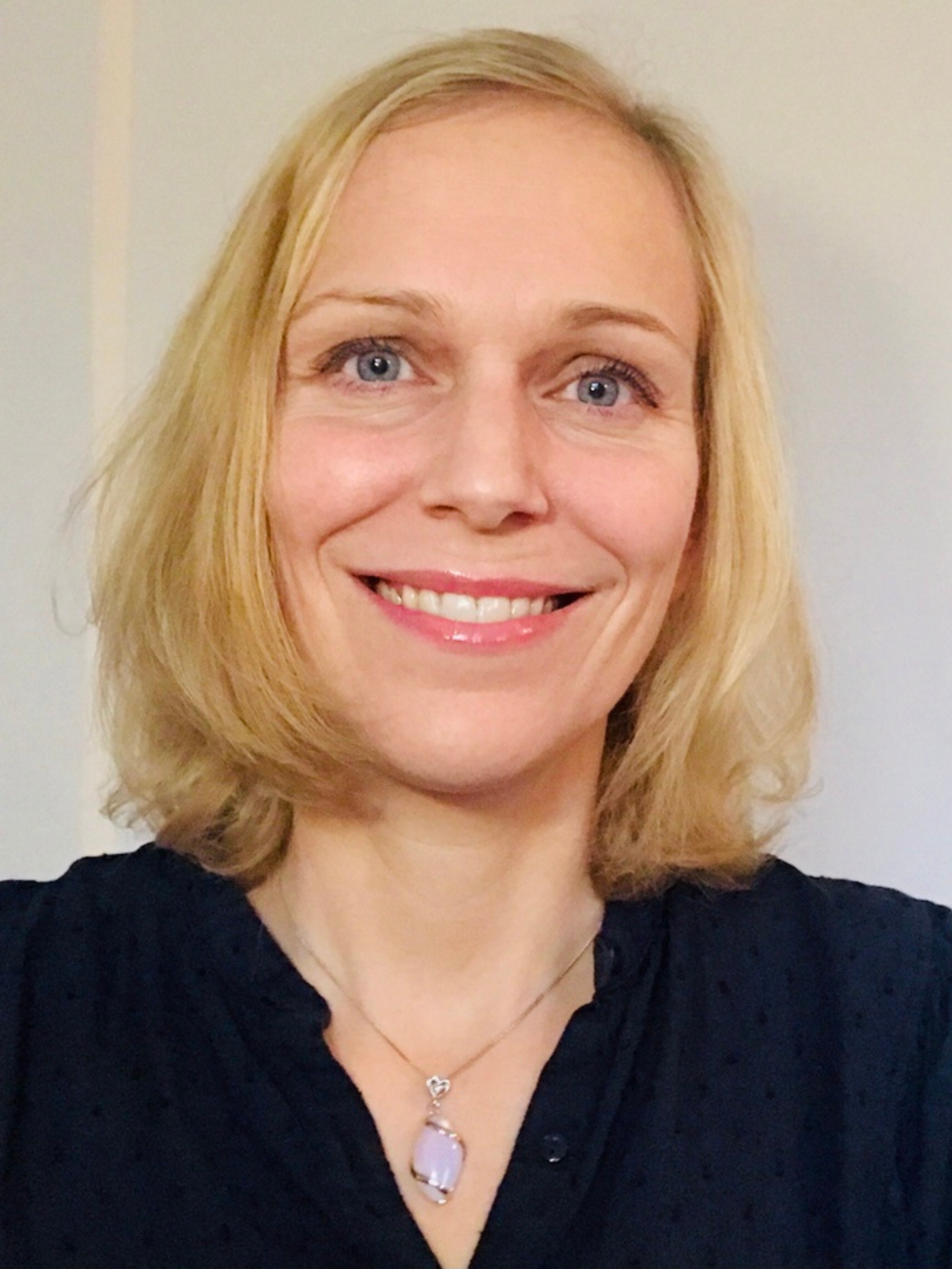 A woman with blonde hair smiling, wearing a black top and a necklace with a purple gemstone pendant.