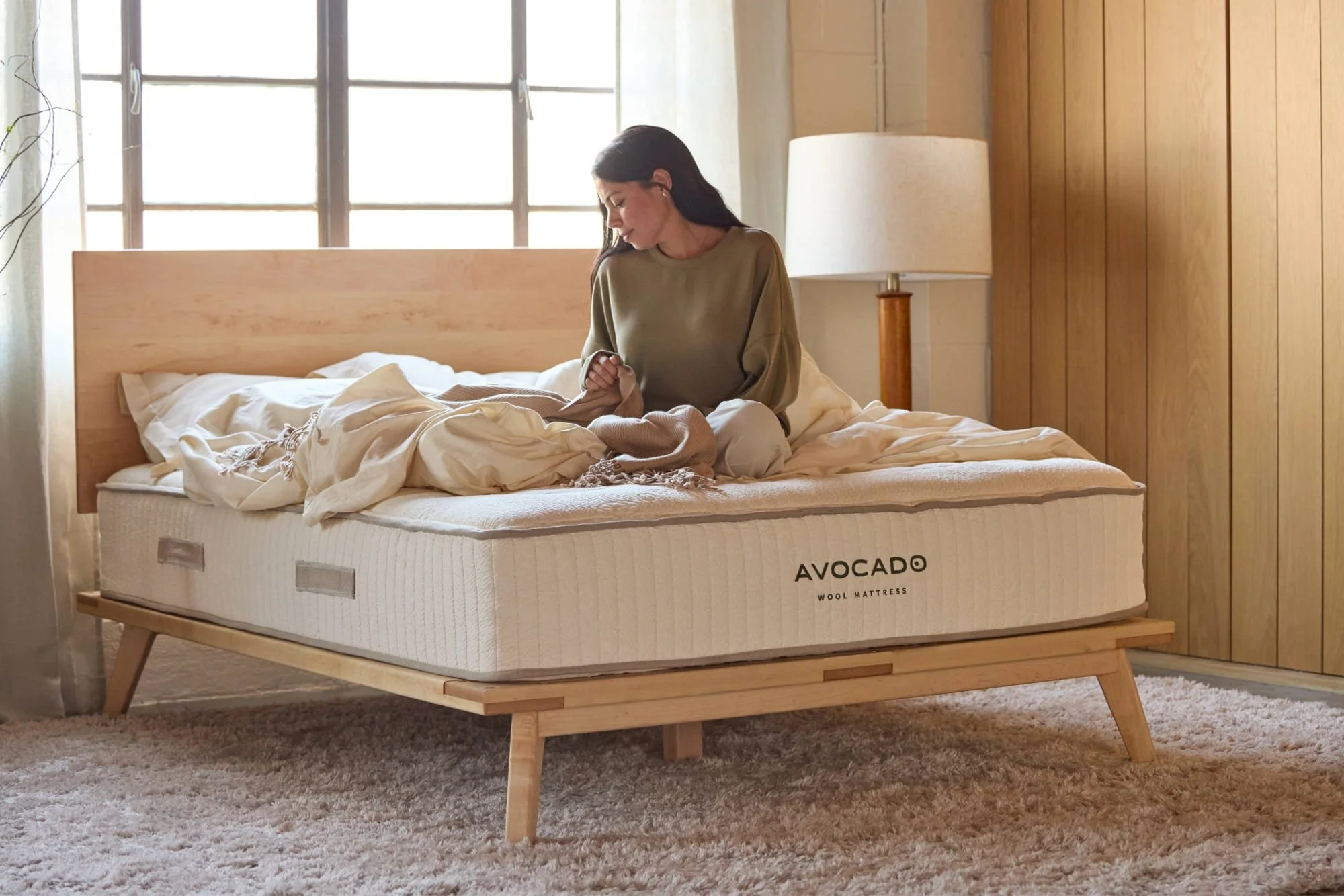 A woman sitting on a mattress in a bedroom with wooden walls and large windows, next to a lamp, with a wool mattress labeled "AVOCADO WOOL MATTRESS".