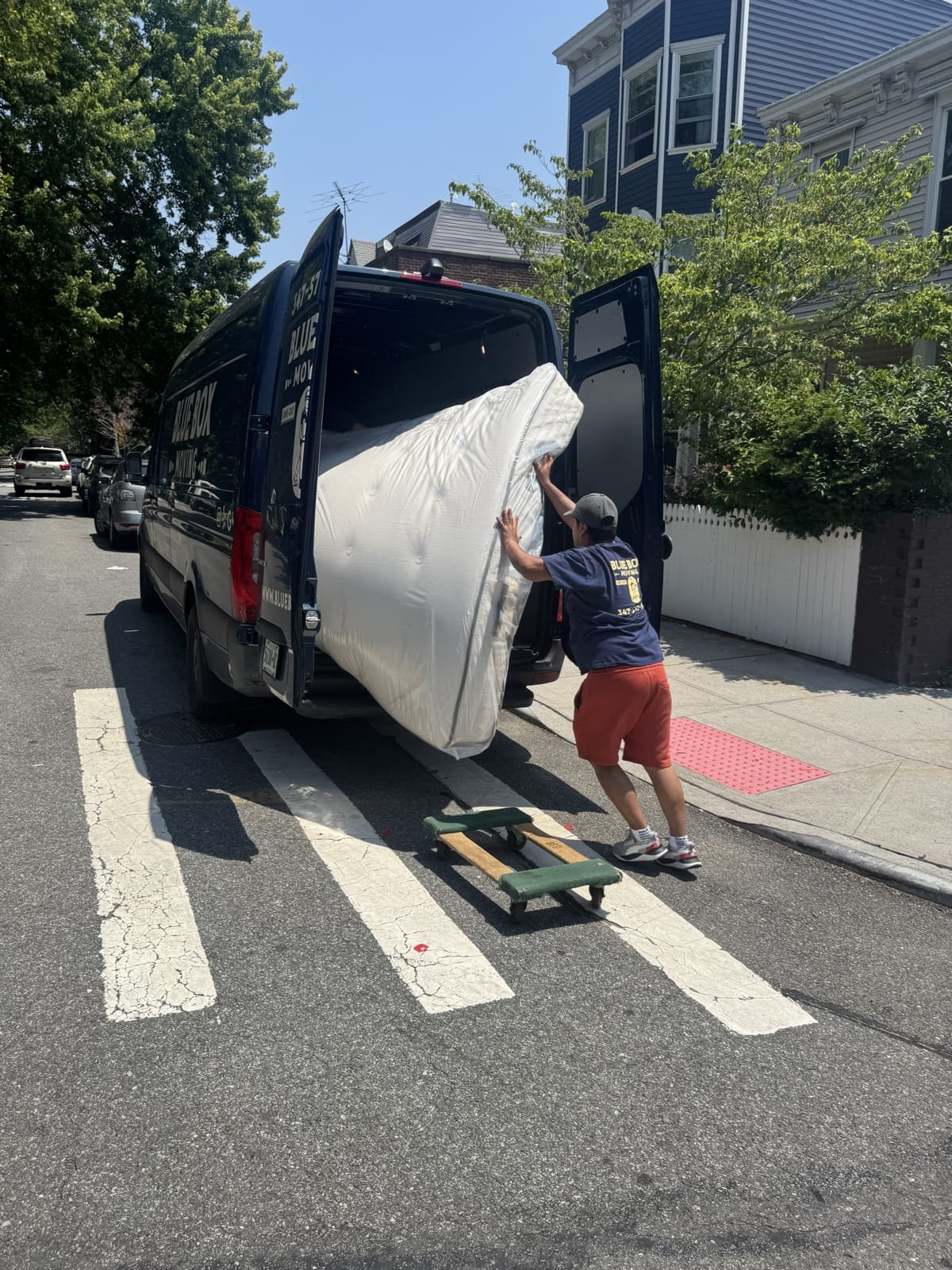 A person loading a mattress into a blue delivery van on a city street on a sunny day.
