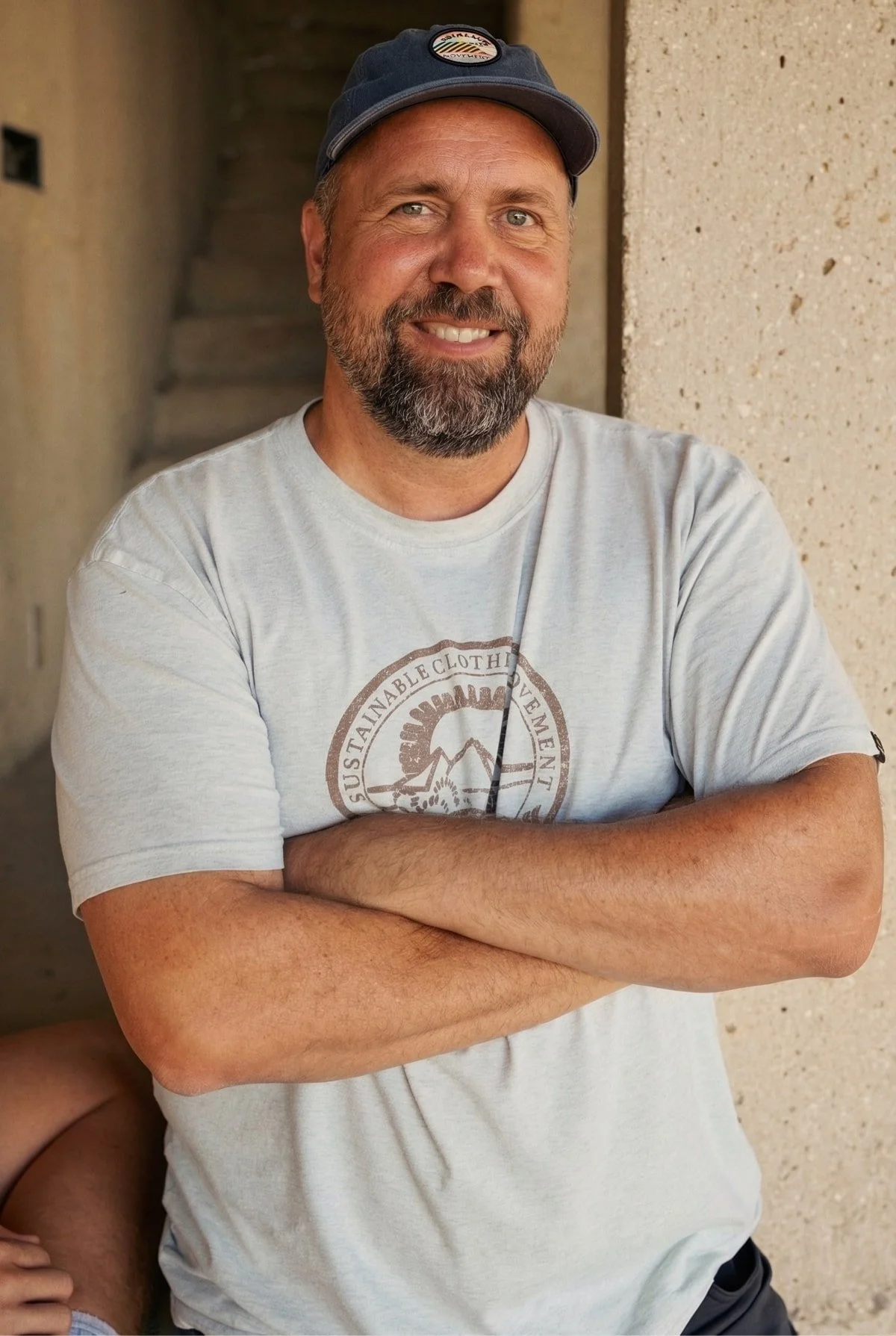A man with a beard and mustache wearing a baseball cap and a light T-shirt standing with crossed arms against a beige wall.