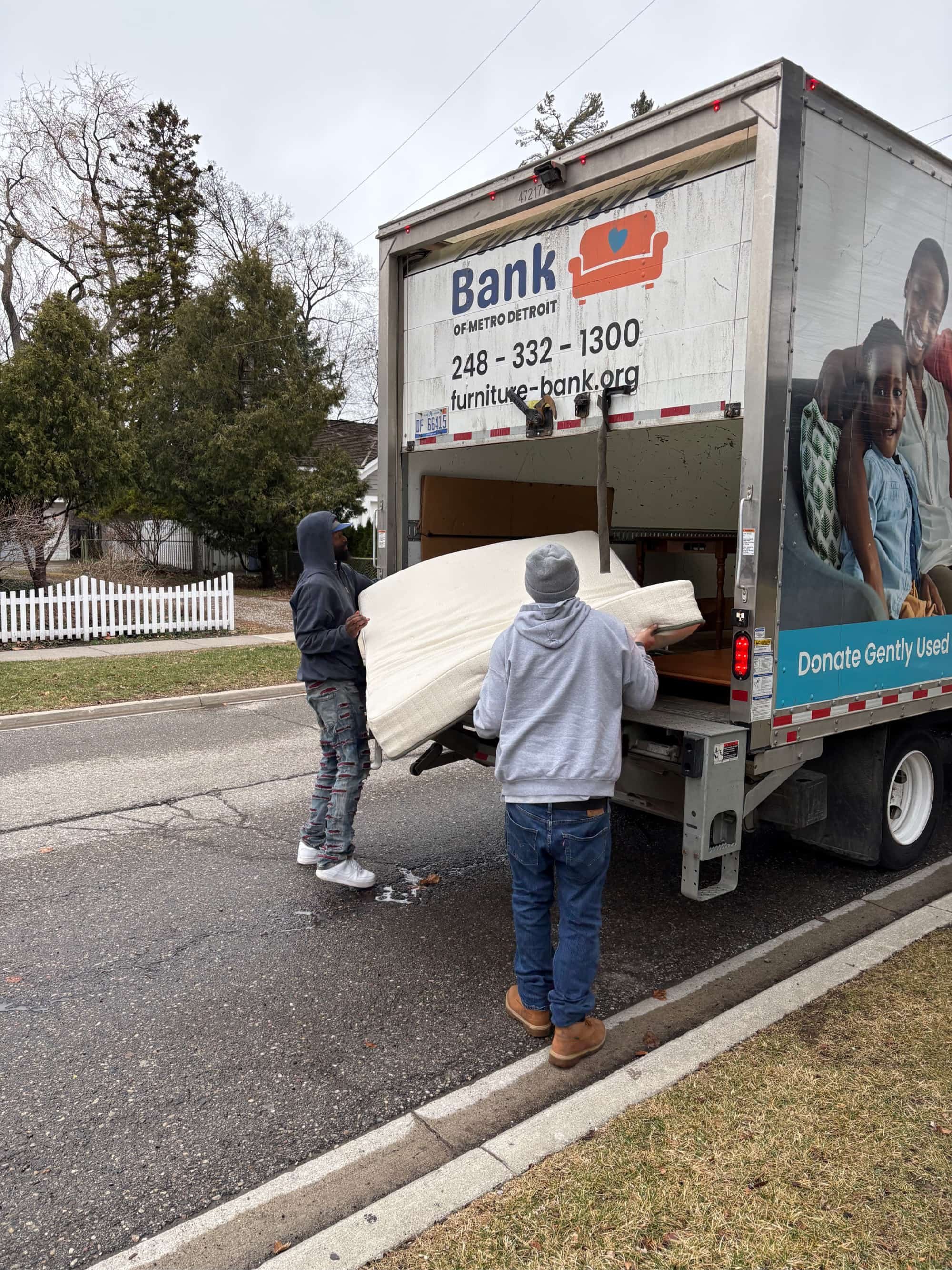 Two people unloading a donated mattress from a truck with a sign for a furniture bank.