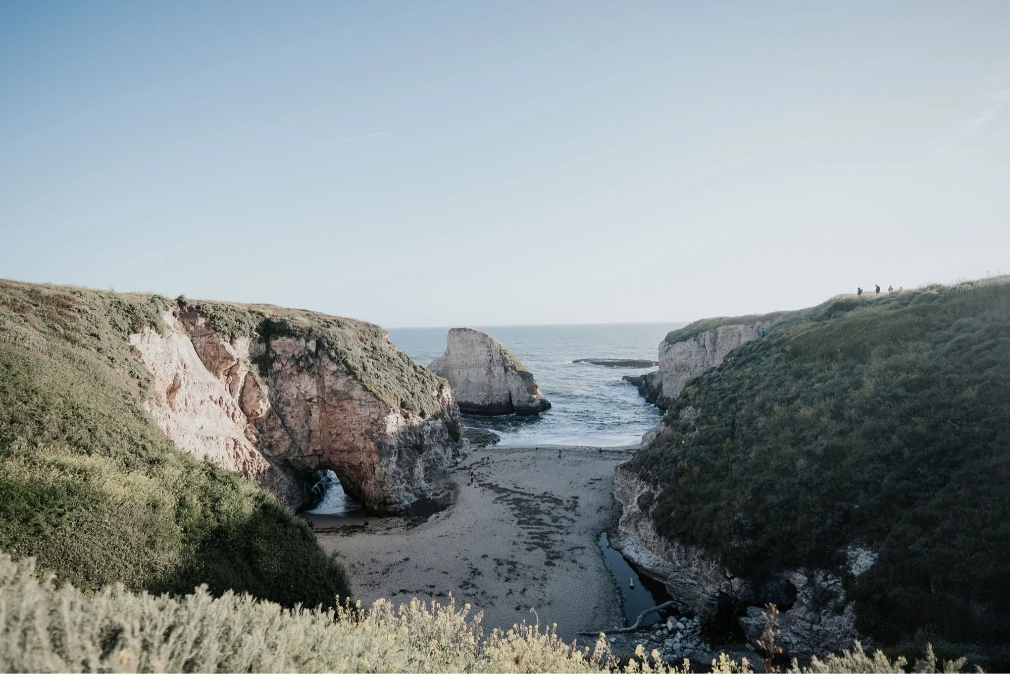 A scenic view of a coastal cove with white cliffs, green vegetation, and a sandy beach, with the ocean in the background and a few small groups of people walking on the cliffs.