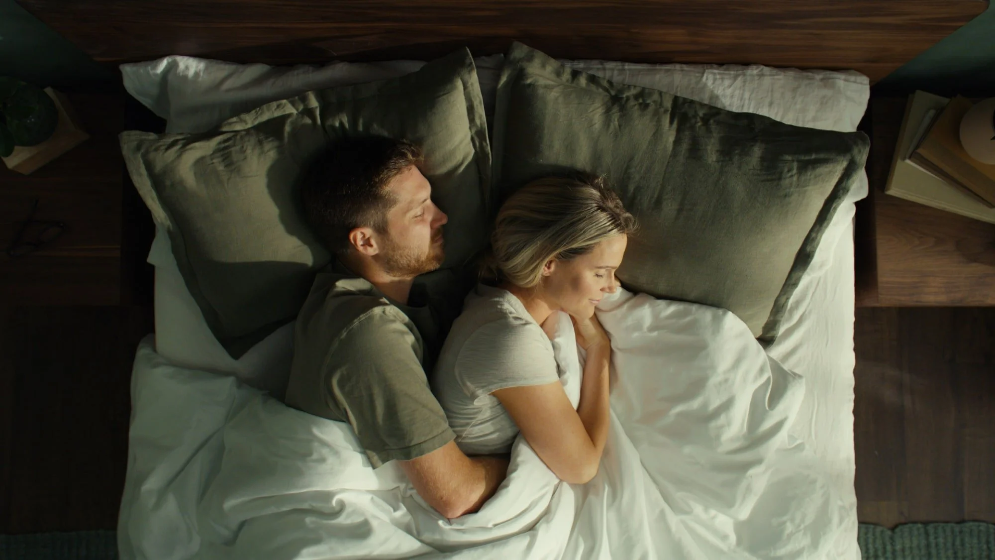A man and a woman sleeping peacefully side by side in bed, facing opposite directions, with their arms resting on each other, covered by a white blanket.