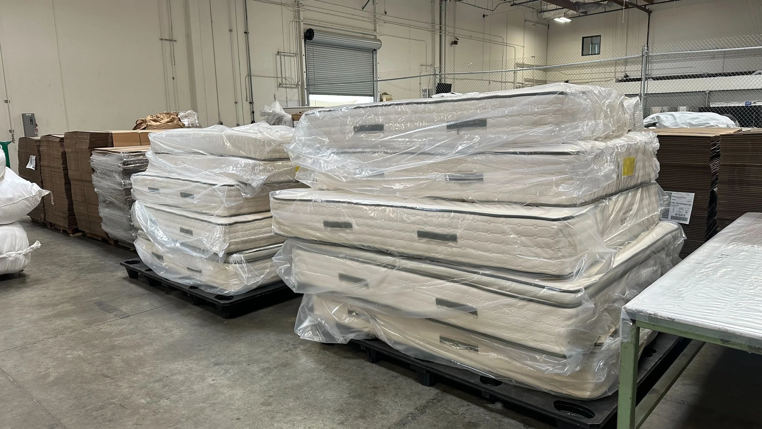 Stacked mattresses wrapped in plastic and cardboard boxes on pallets in a warehouse.