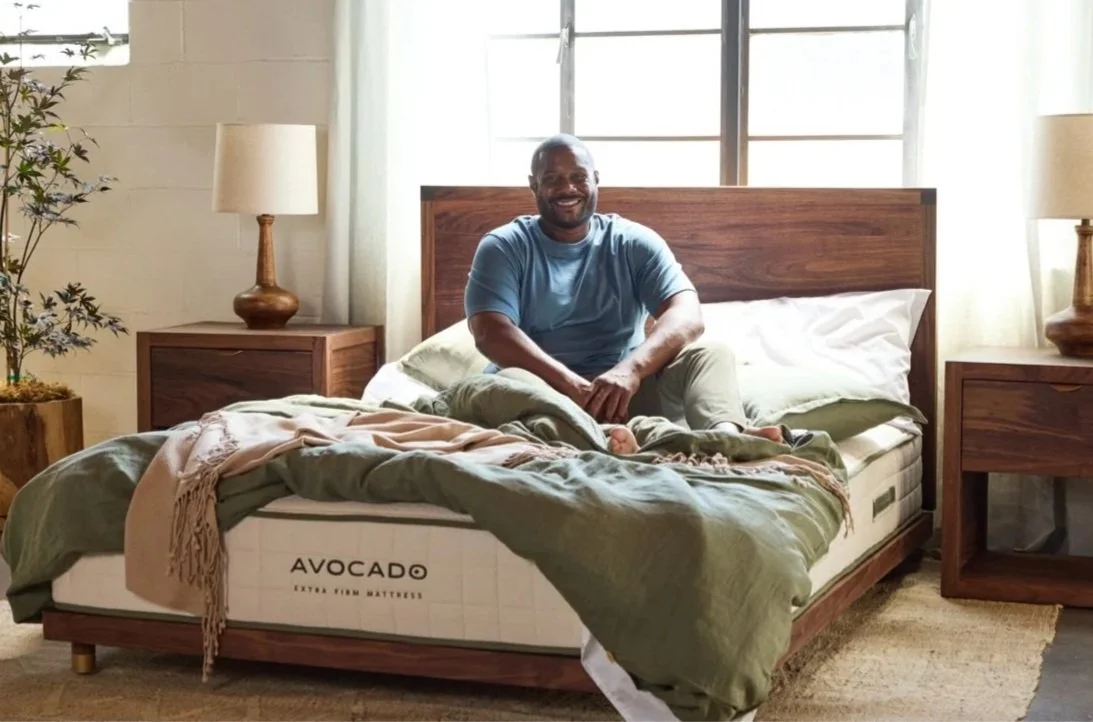 A man sitting on an Avocado mattress in a bedroom with wooden nightstands and lamps. Large windows behind the bed let in natural light.
