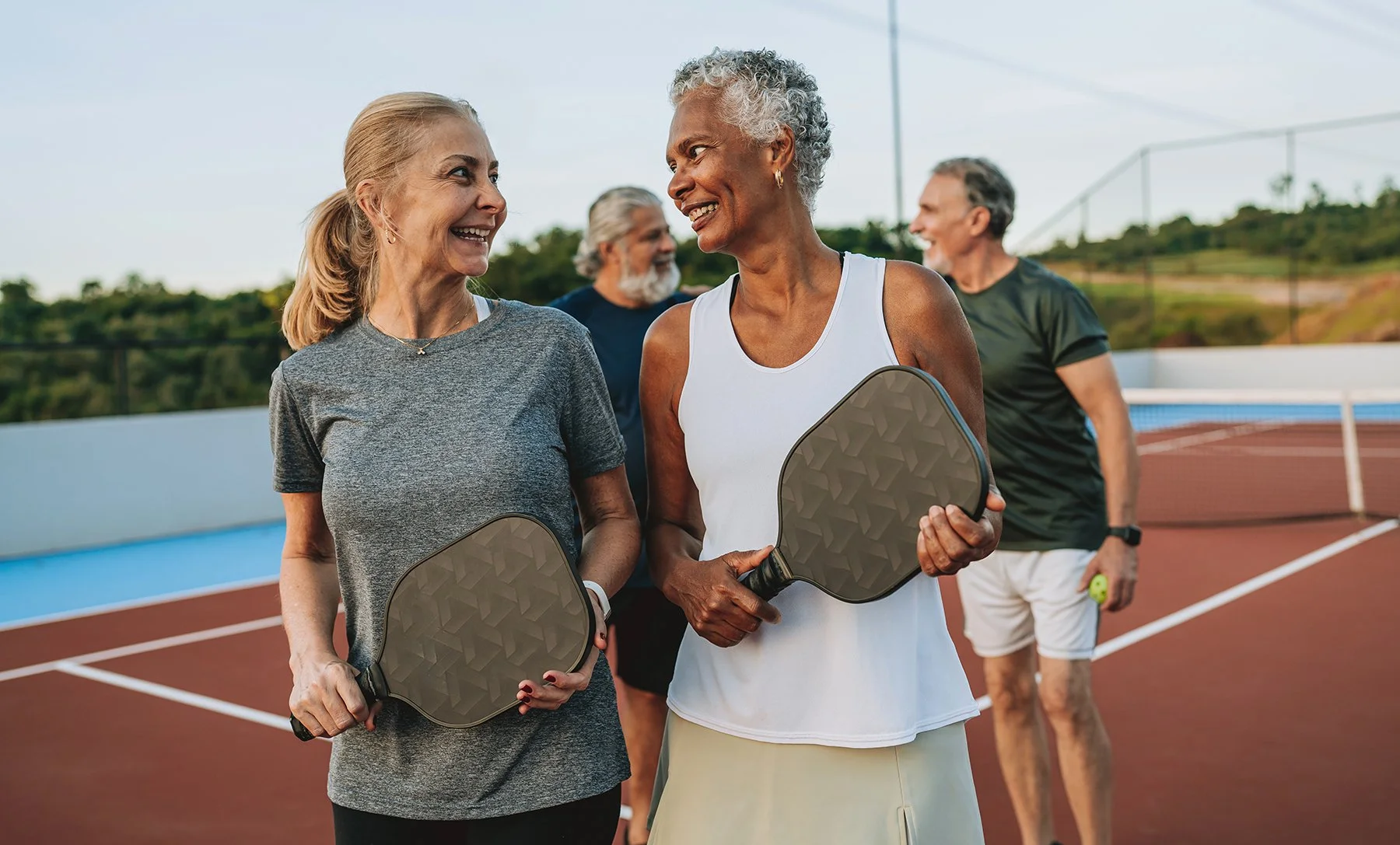 Four older adults standing on a tennis court with two women in the foreground holding paddles and two men in the background. The women are smiling and engaging in conversation.
