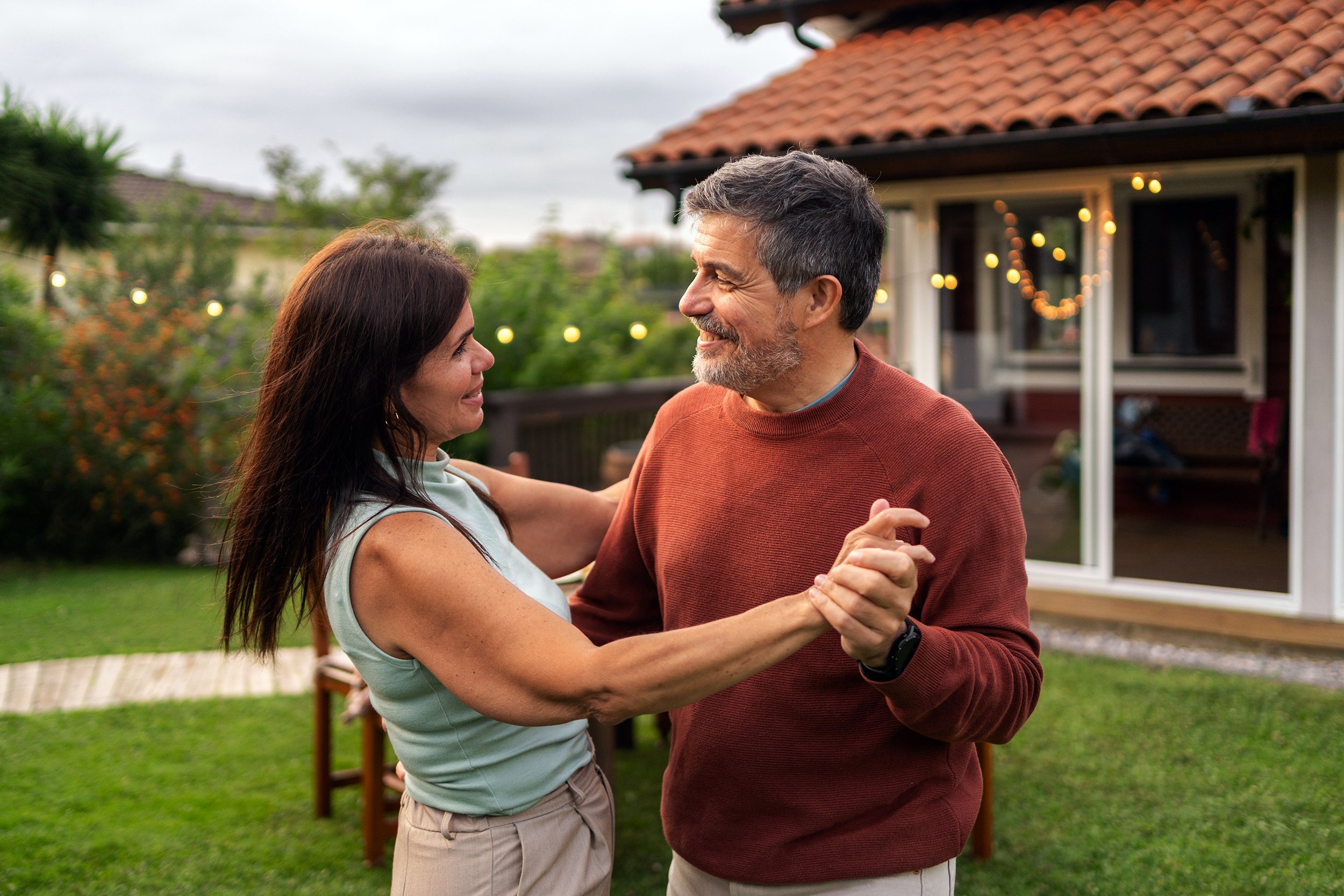 A middle-aged man and woman dance together in their backyard, smiling at each other.