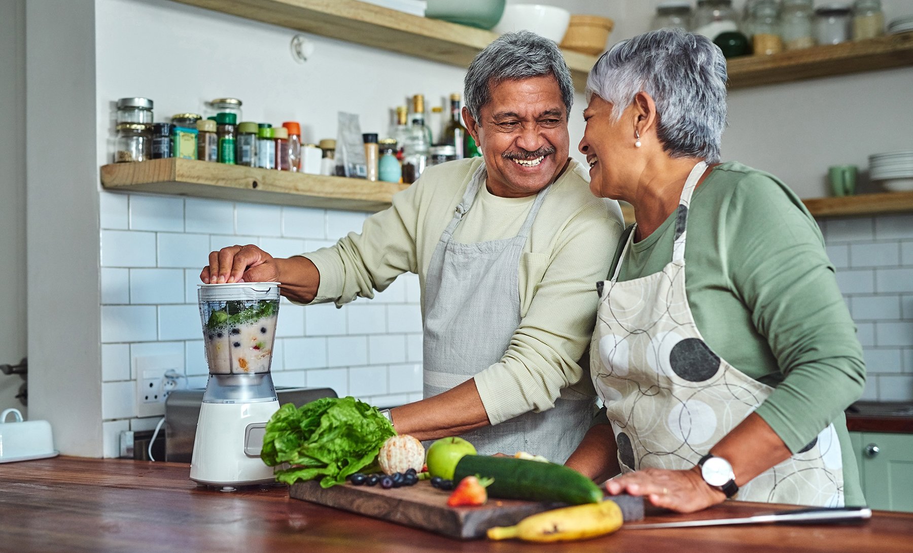 An elderly couple smiling and preparing a smoothie in a kitchen, surrounded by fresh fruits and vegetables.
