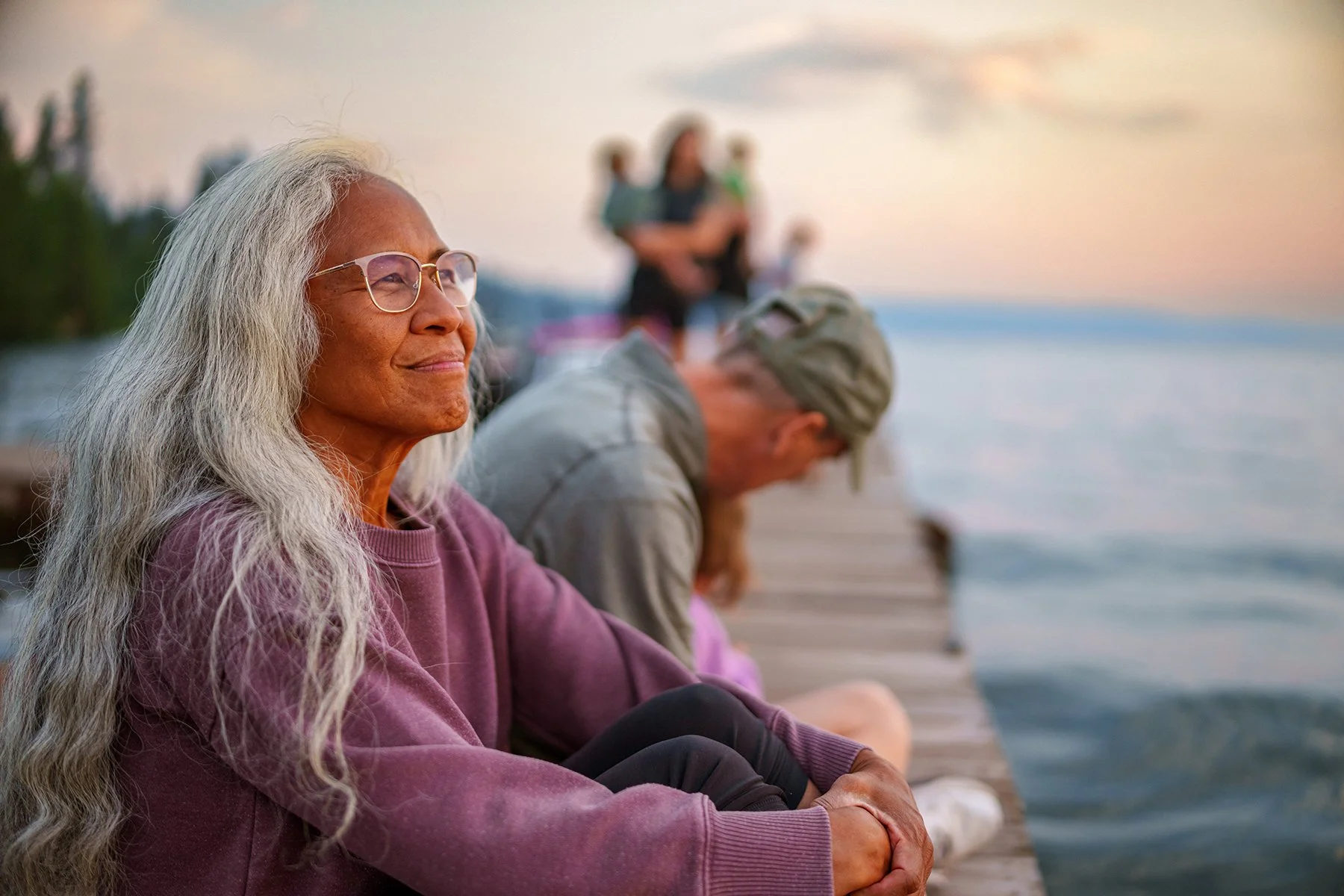 Senior woman with long gray hair and glasses sitting on a dock by the water, smiling and relaxing at sunset, with other people in the background.