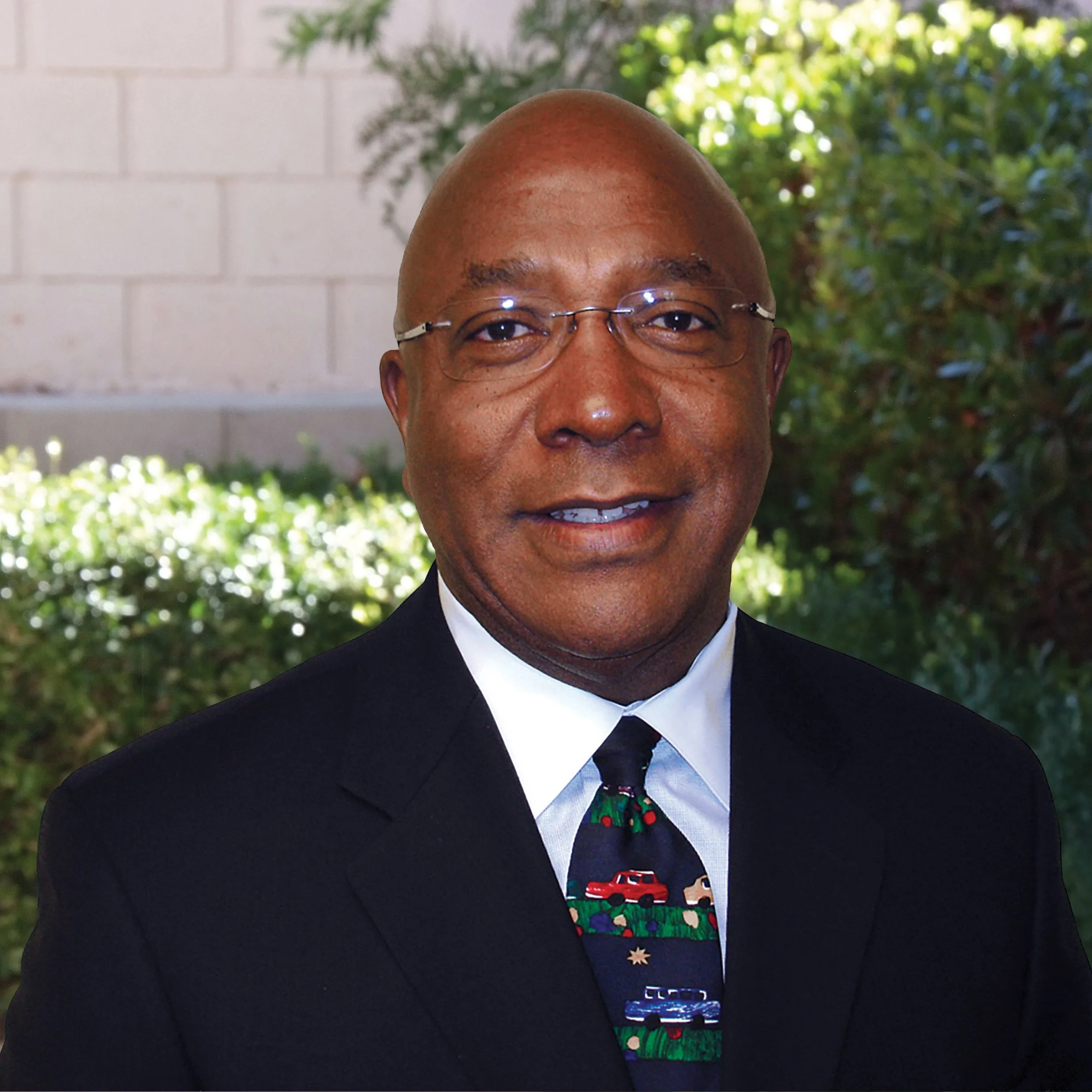 George Wheeler Jr, Founder and Financial Strategist, in a black suit and a colorful tie, standing outdoors with green foliage and a white brick wall in the background.