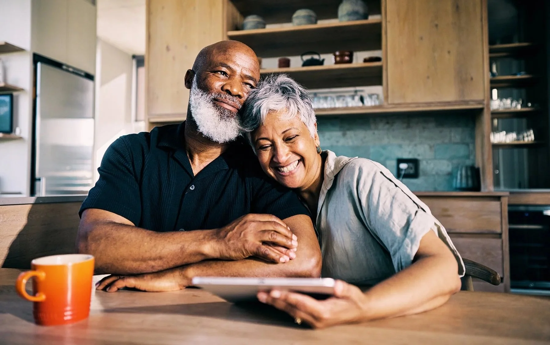 A happy elderly couple with gray hair, sharing an affectionate moment at the kitchen table, looking at a tablet.