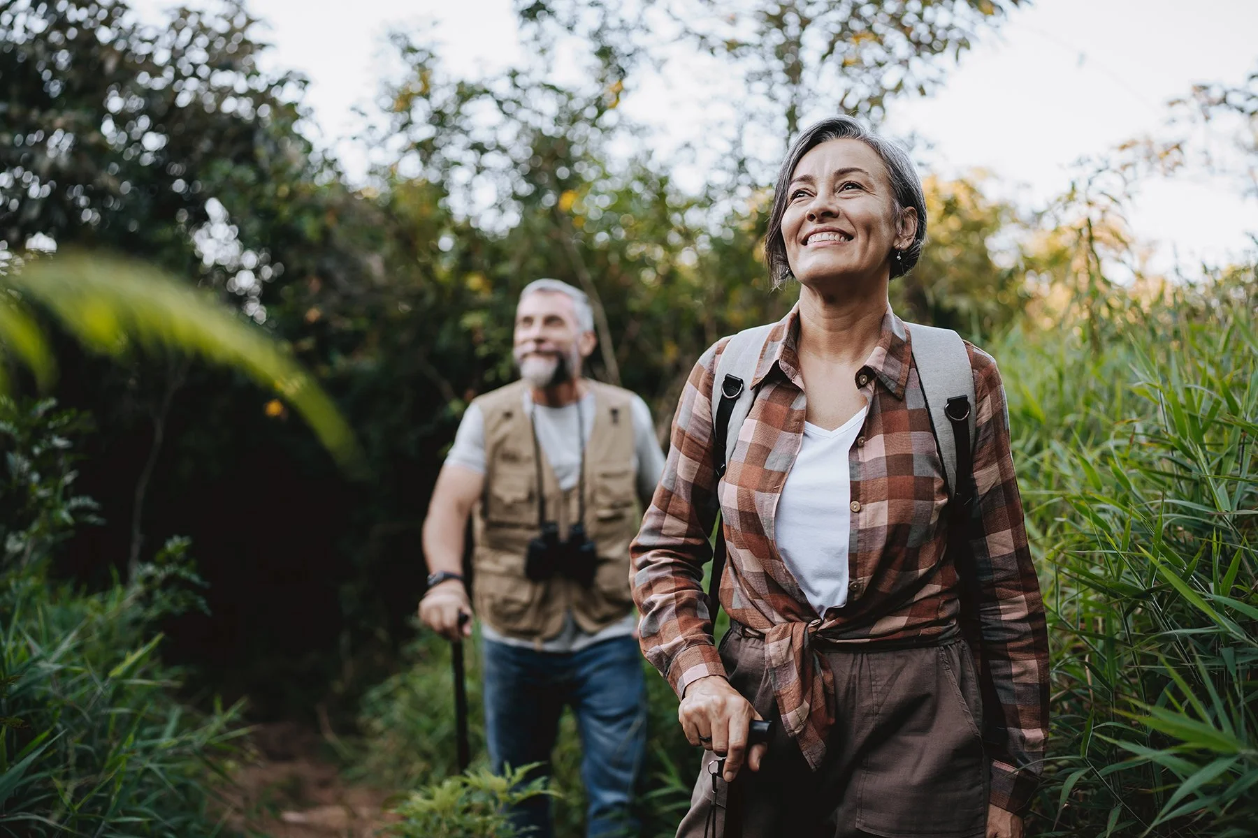 A smiling woman and a man hiking through a lush, green forest. The woman in the foreground wears plaid shirt and carries a backpack, while the man in the background wears a vest and also carries a backpack.