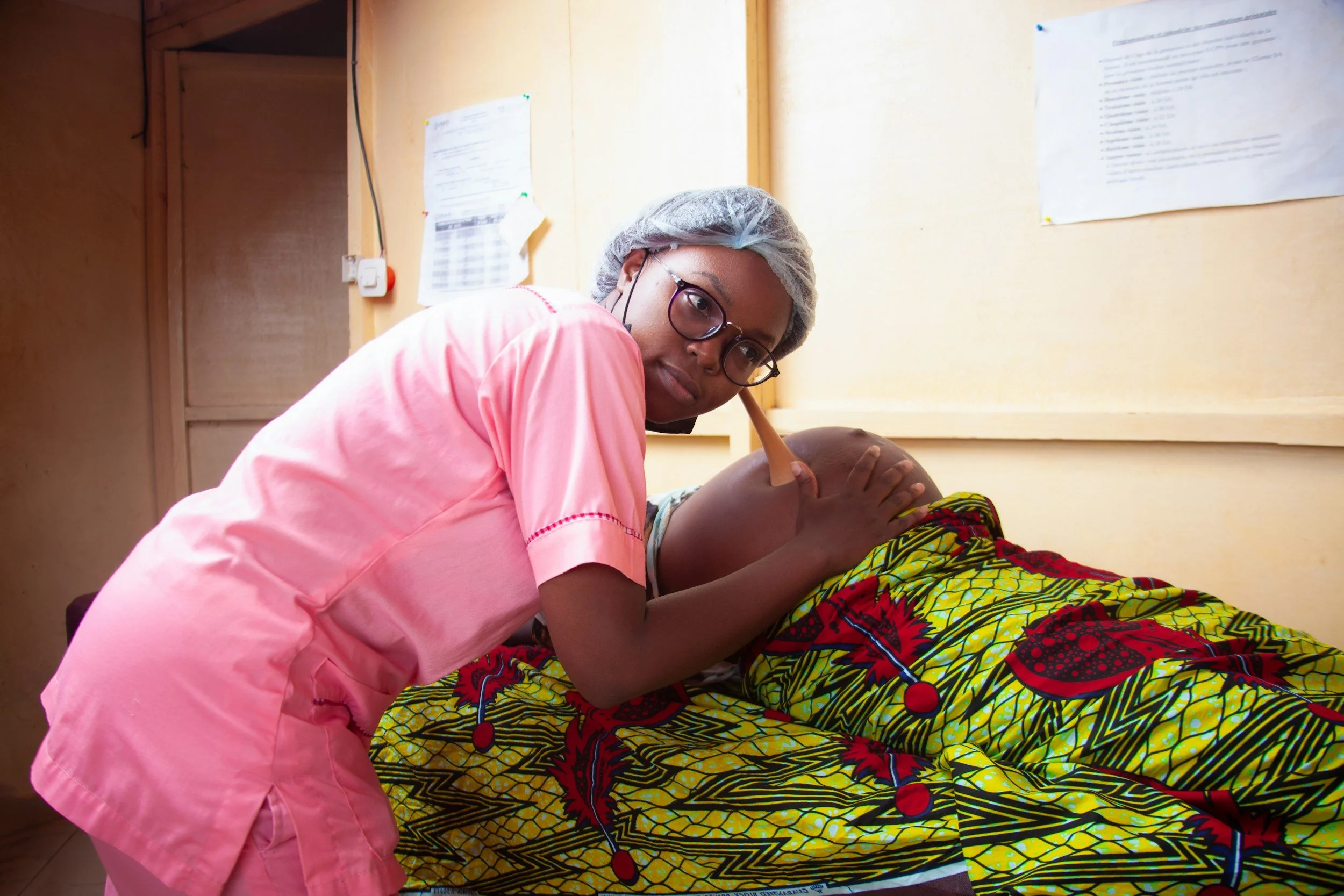 A healthcare worker giving a medical emergency training or massage to a patient lying on a bed, in a room with light-colored walls and documents on the wall.
