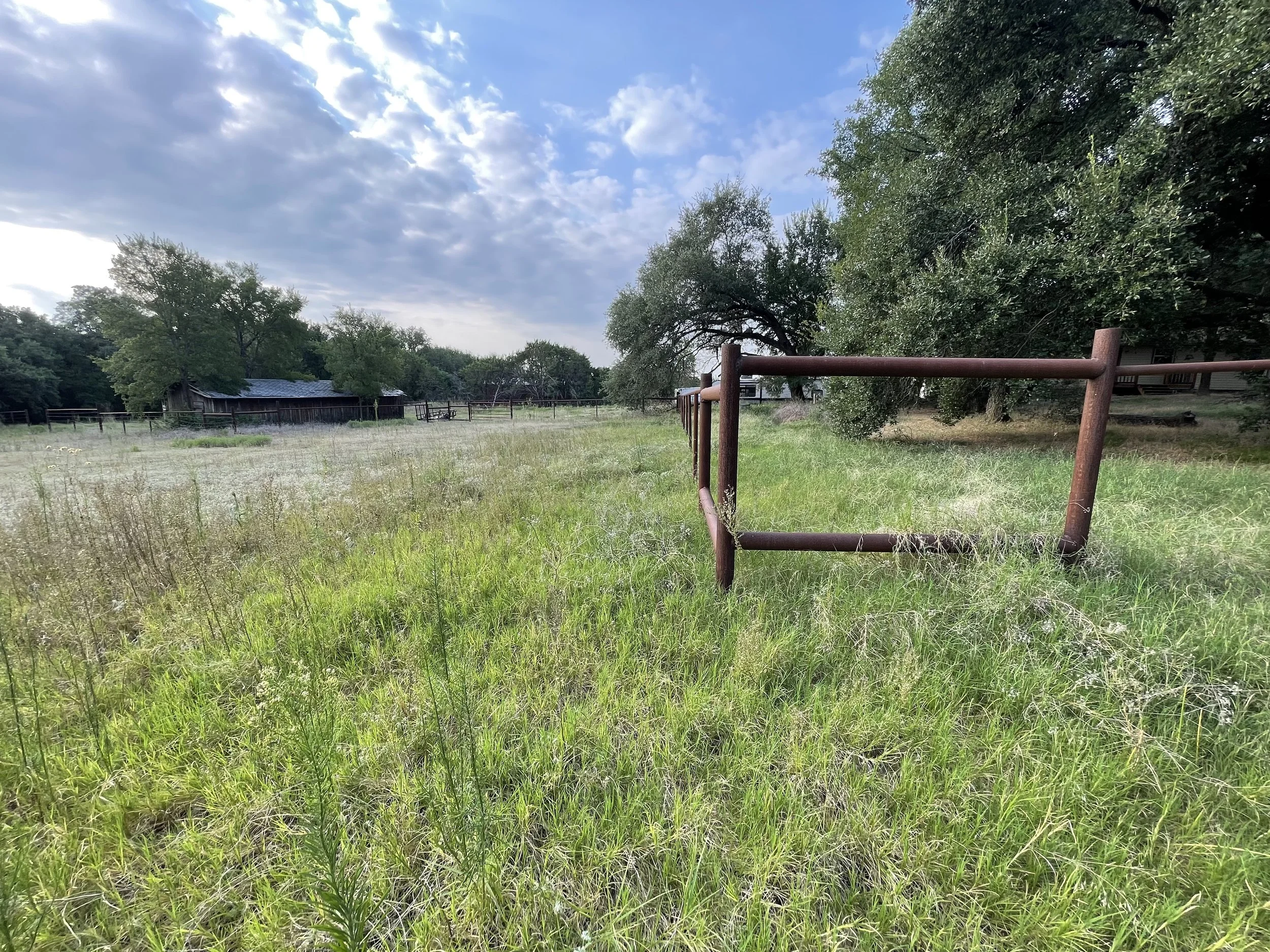 A rural scene with green grass, trees, a small weathered barn, and a rusty fence, under a partly cloudy sky.