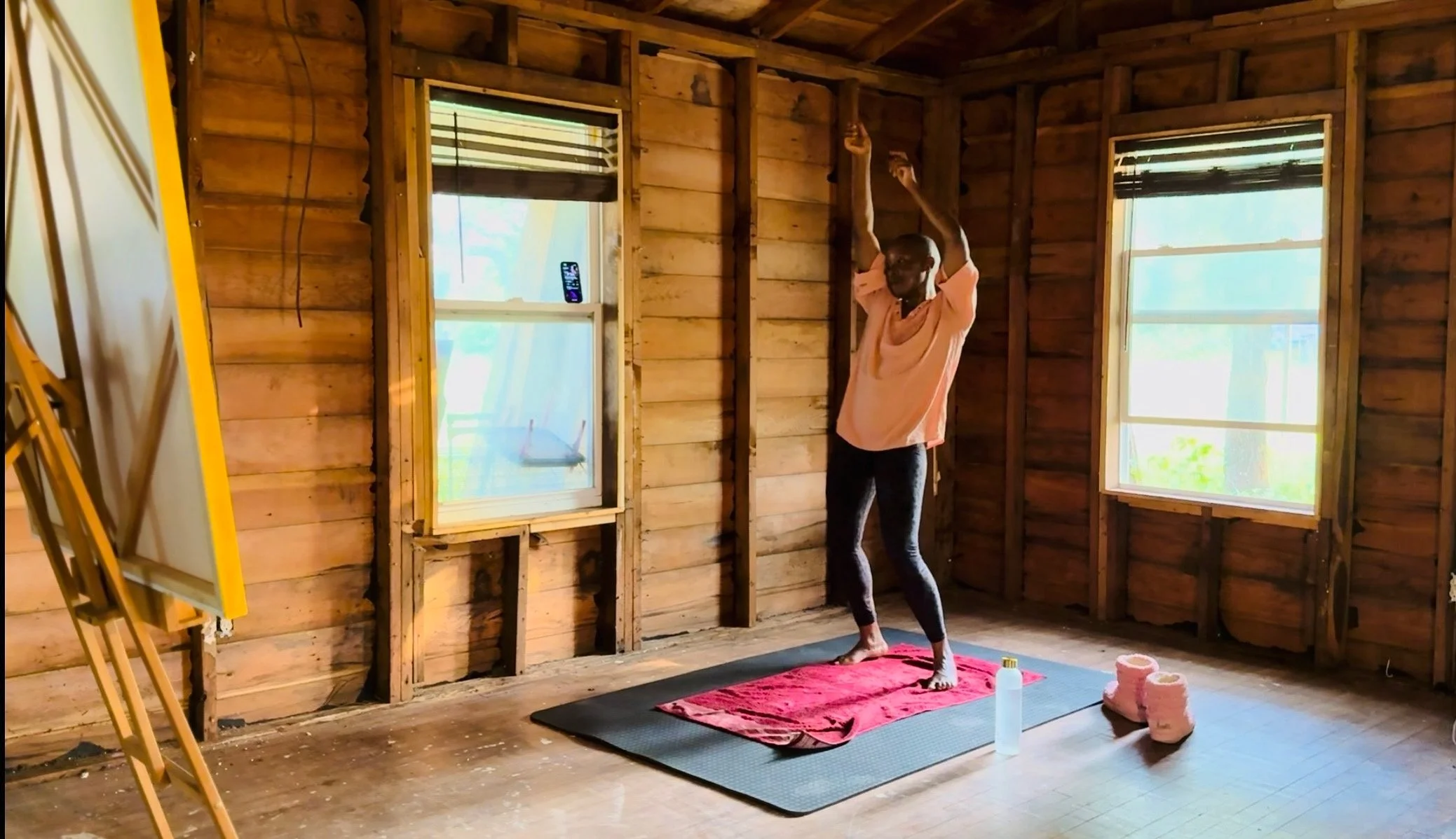 A person practicing yoga in a partially constructed wooden room with two windows, a yoga mat, pink towels, and a water bottle.