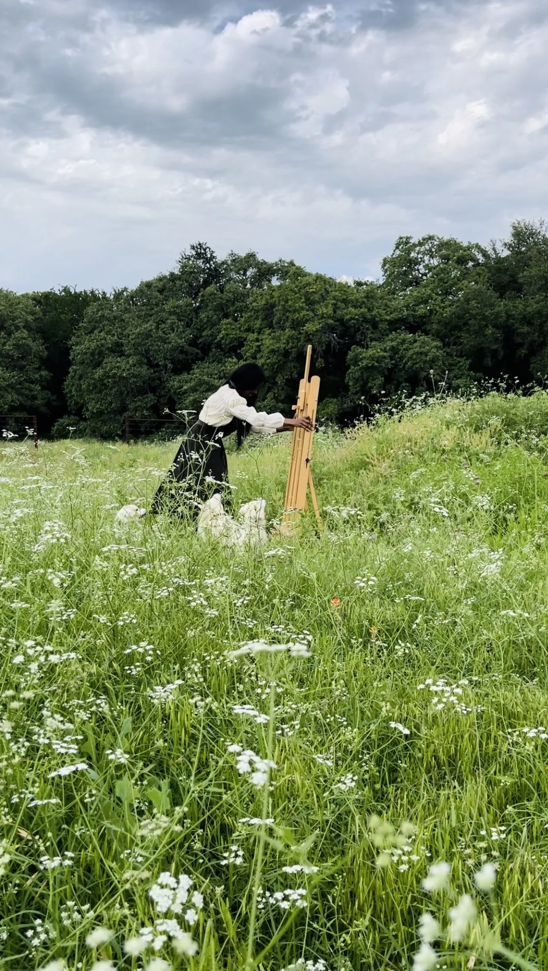 Person in white shirt and black skirt painting outdoors on an easel in a grassy field with wildflowers, surrounded by trees under a cloudy sky.