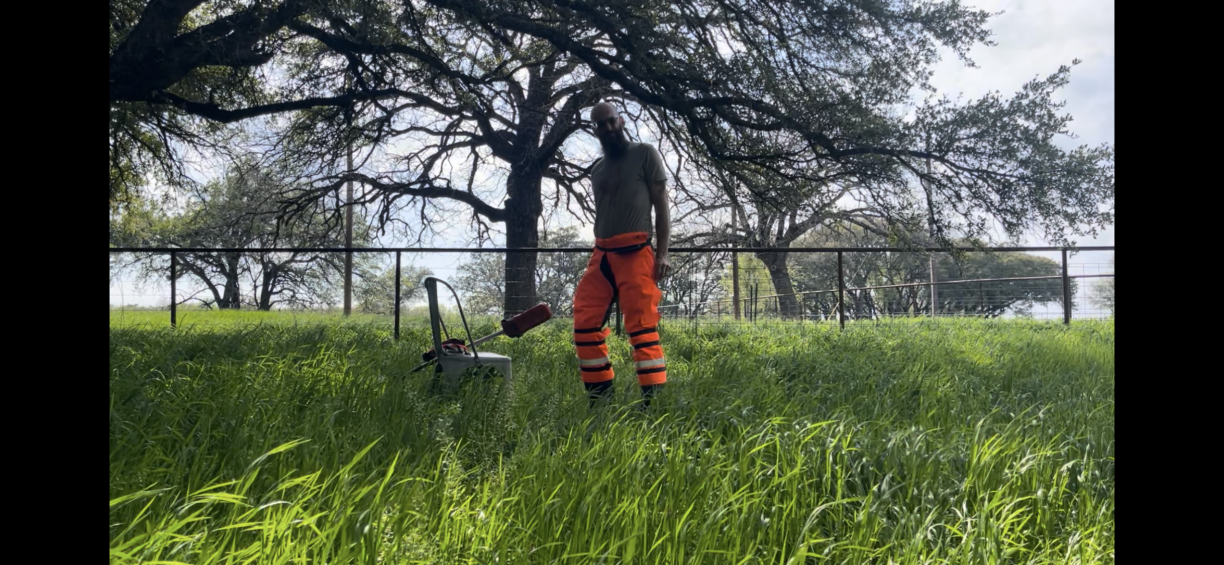 A man wearing orange and black work pants, a gray t-shirt, and sunglasses standing outdoors on green grass, with a lawnmower nearby. Behind him, there is a large tree with many branches, some with leaves and some without, and a fence in the backgroun