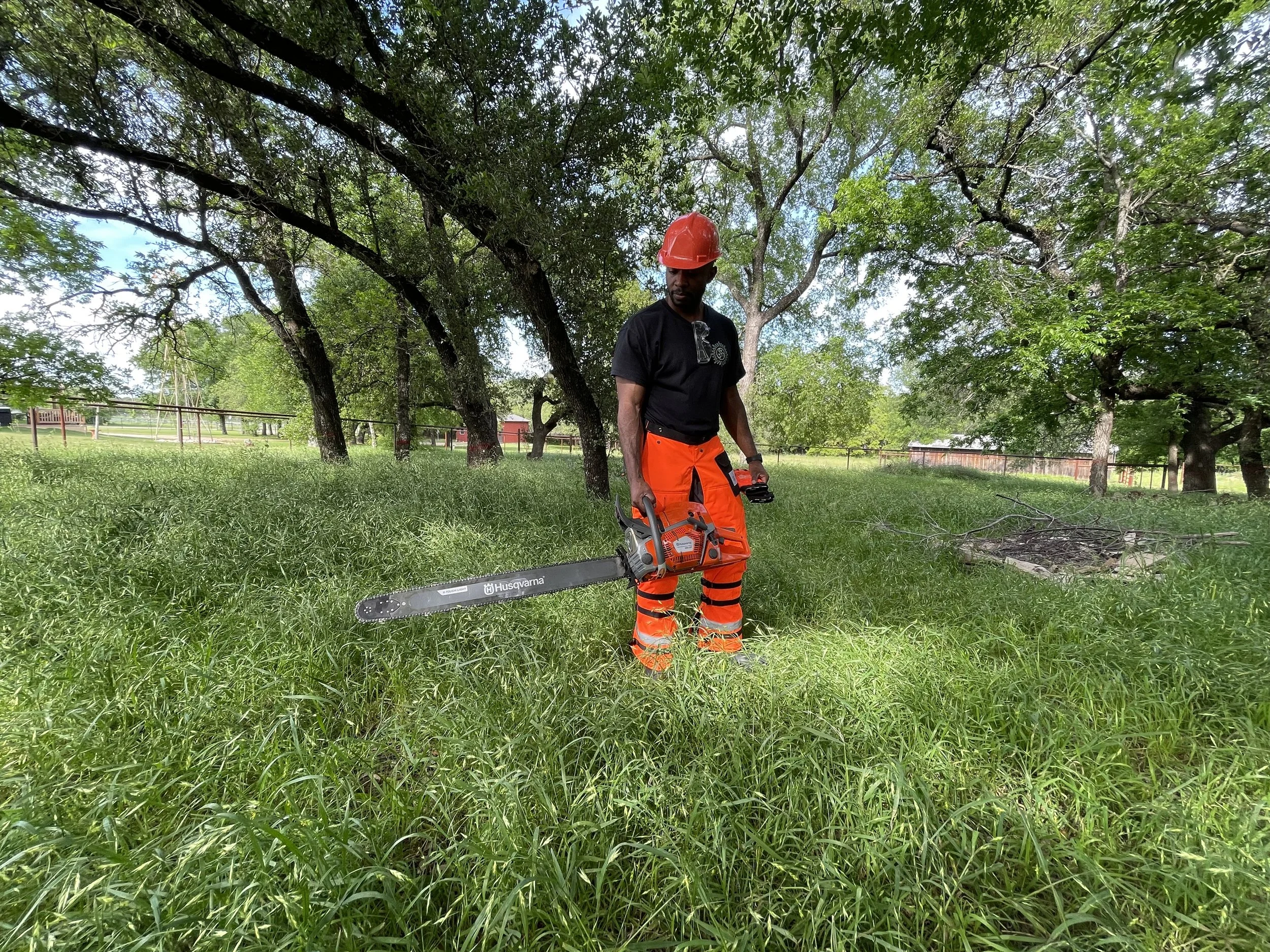 A man wearing an orange hard hat and orange work pants is outdoors, holding a chainsaw, surrounded by green grass and trees.