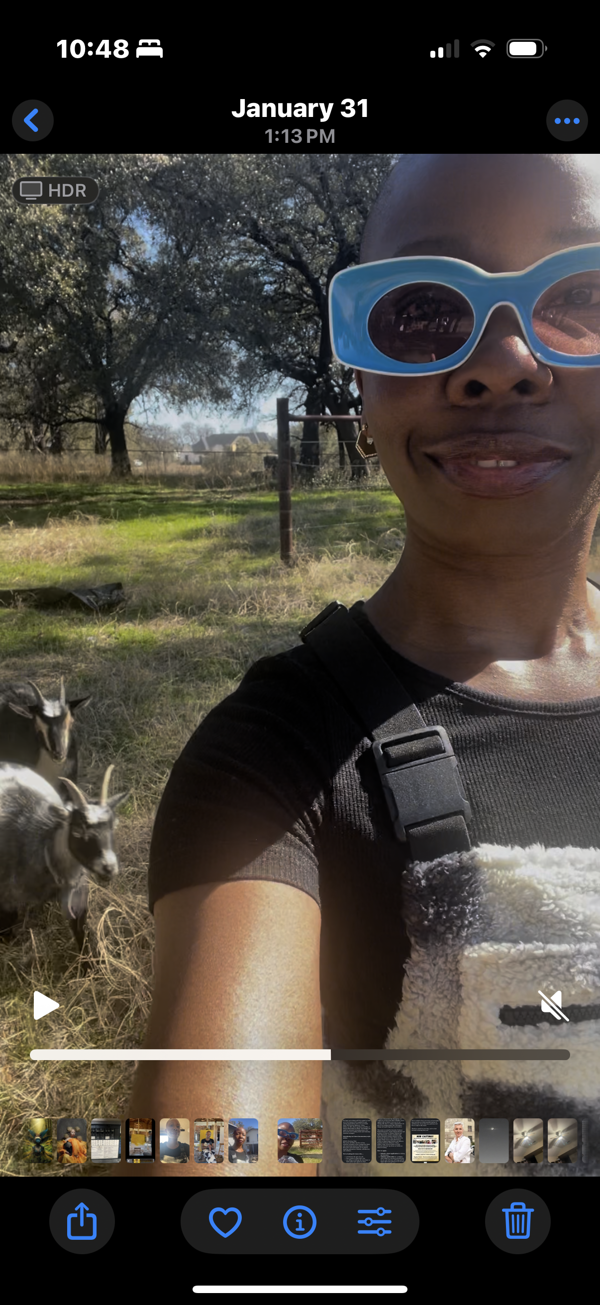 A woman taking a selfie outdoors wearing large blue sunglasses and a black shirt, with goats standing on dry grass behind her and trees in the background.