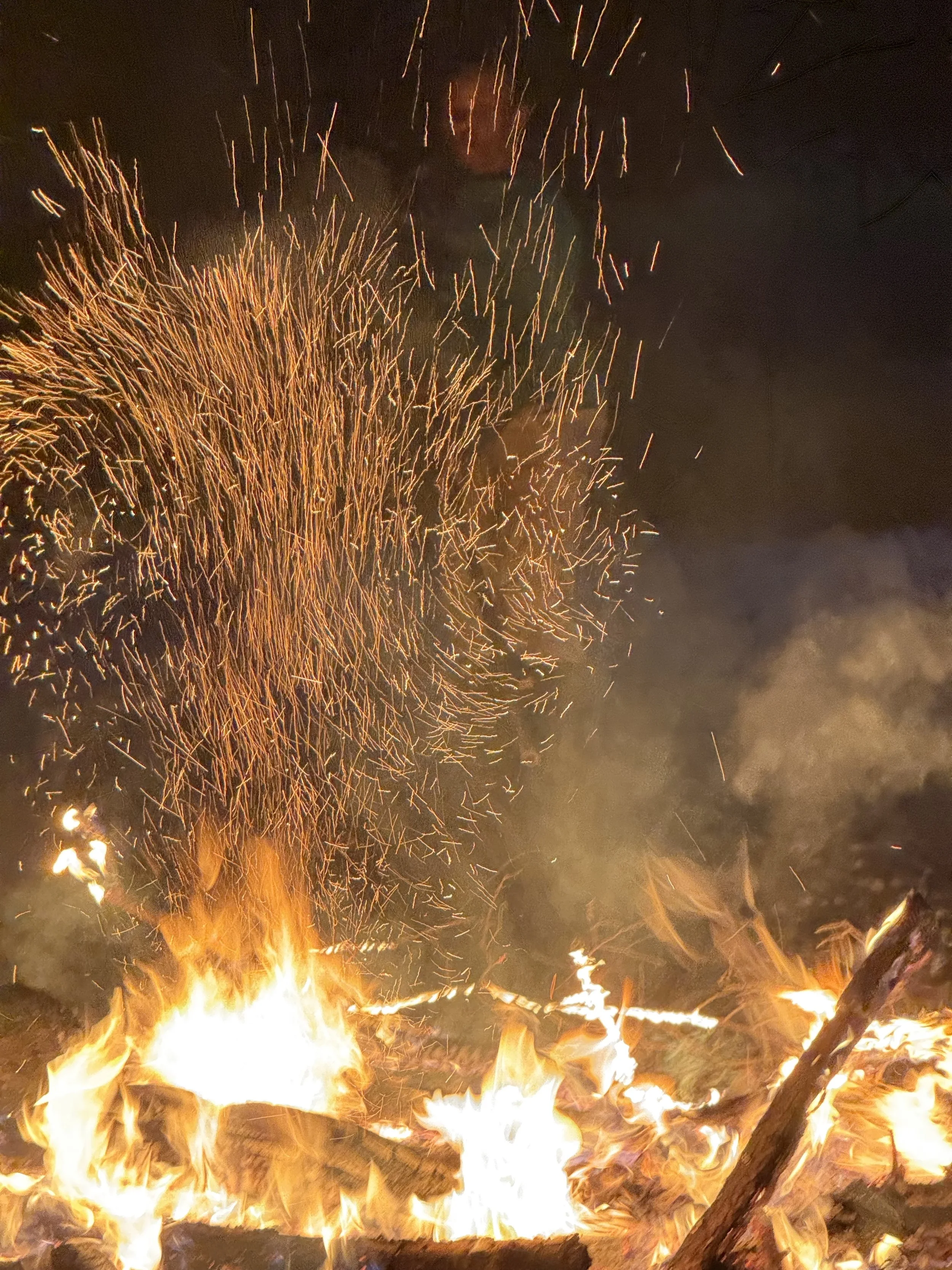 A large outdoor fire with bright flames at the bottom and fireworks exploding in the night sky above.