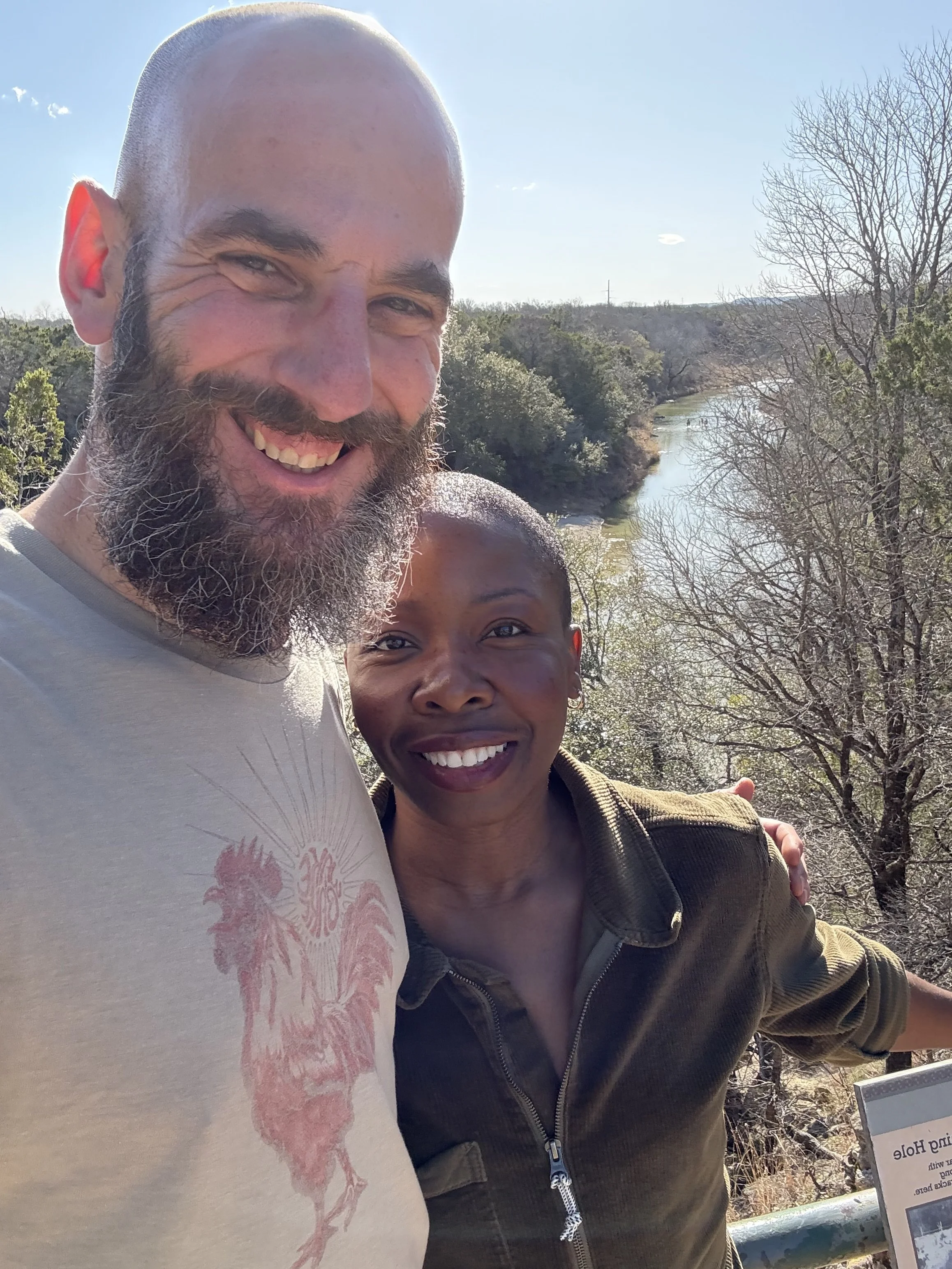 A smiling man with a beard and a bald head taking a selfie outdoors near a river and trees, with a partly cloudy sky.