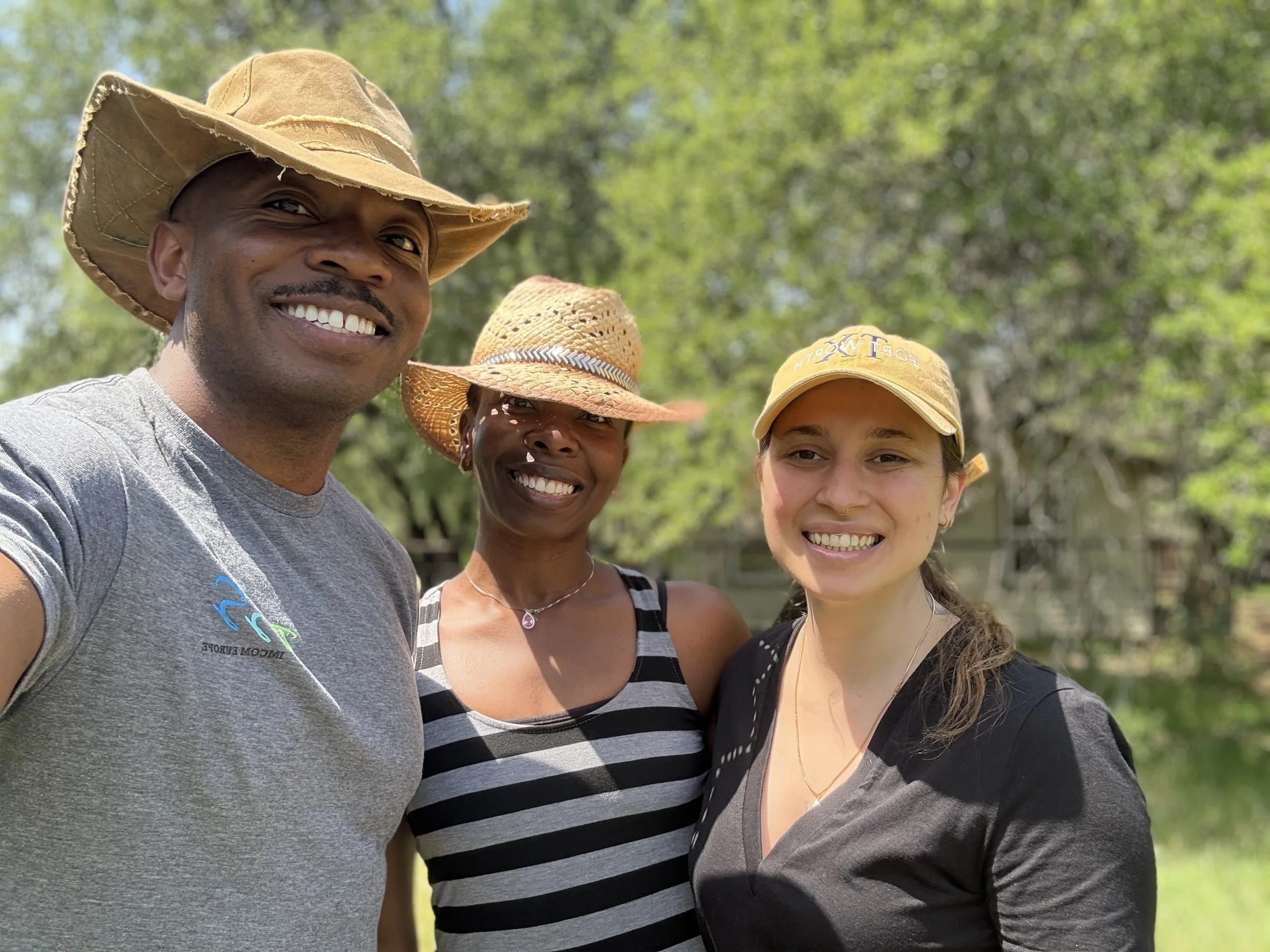 Three smiling people, two women and one man, taking a selfie outdoors with green trees in the background. They are wearing casual clothes and hats.