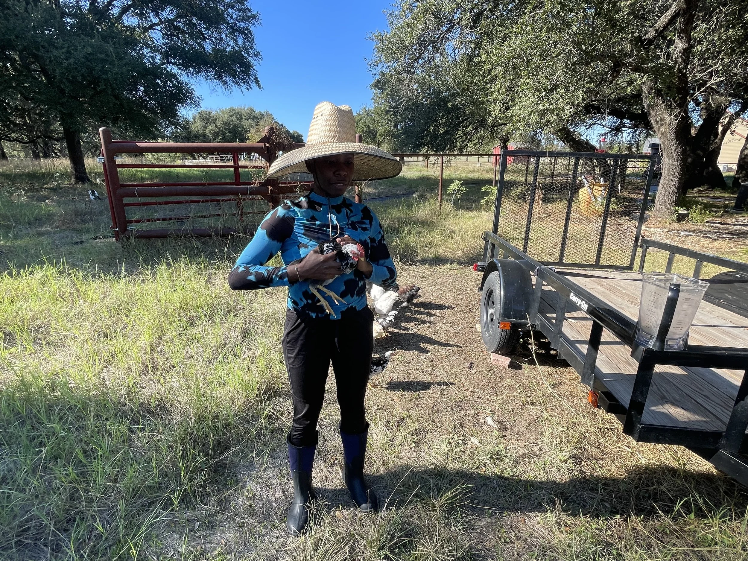 A woman wearing a large straw hat and a blue cycling jersey standing outdoors near a trailer, holding a rooster, with trees, fencing, and a clear blue sky in the background.