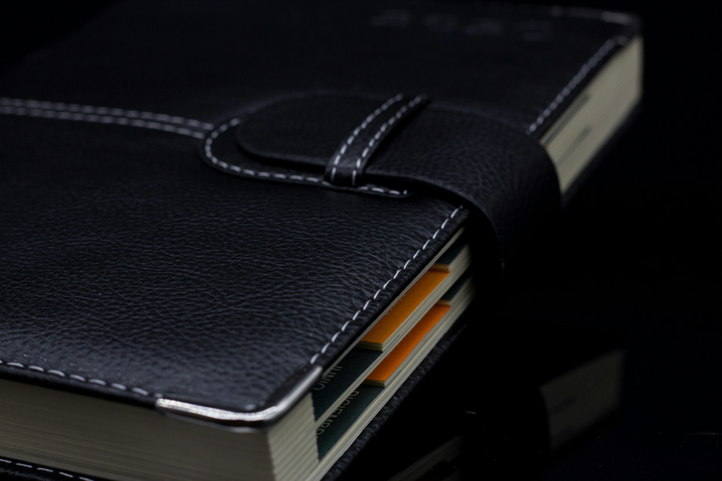 A black leather-bound planner or notebook with white stitching, slightly open to reveal some colorful pages inside, on a dark surface.